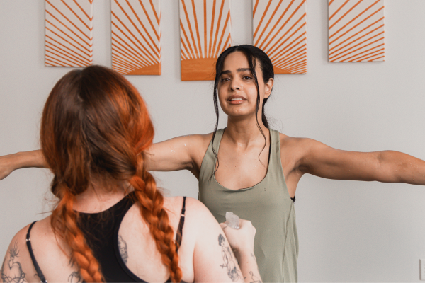 Two women with tattoos, one with red hair and the other with dark hair and a green top, appear to be practicing boxing or a fitness workout indoors, with artwork hanging on the wall behind them.