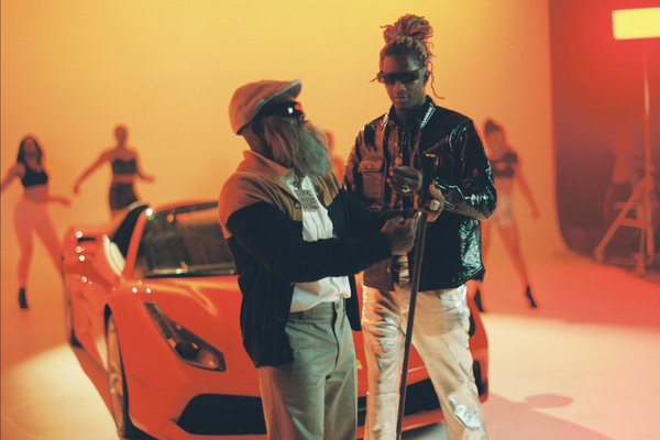 Two men in stylish clothing and sunglasses standing near a yellow sports car, with a woman in a dance pose blurred in the background, on a brightly lit set with warm tones.
