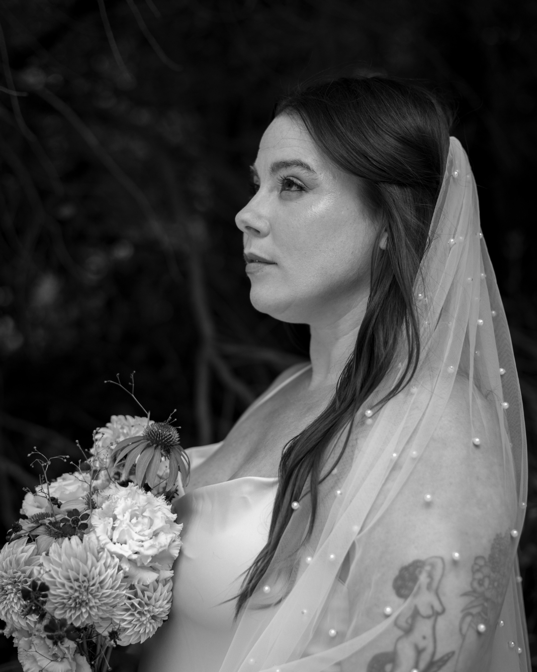 Black and white photo of a woman with a veil, holding a bouquet of flowers, with tattoos visible on her arm.