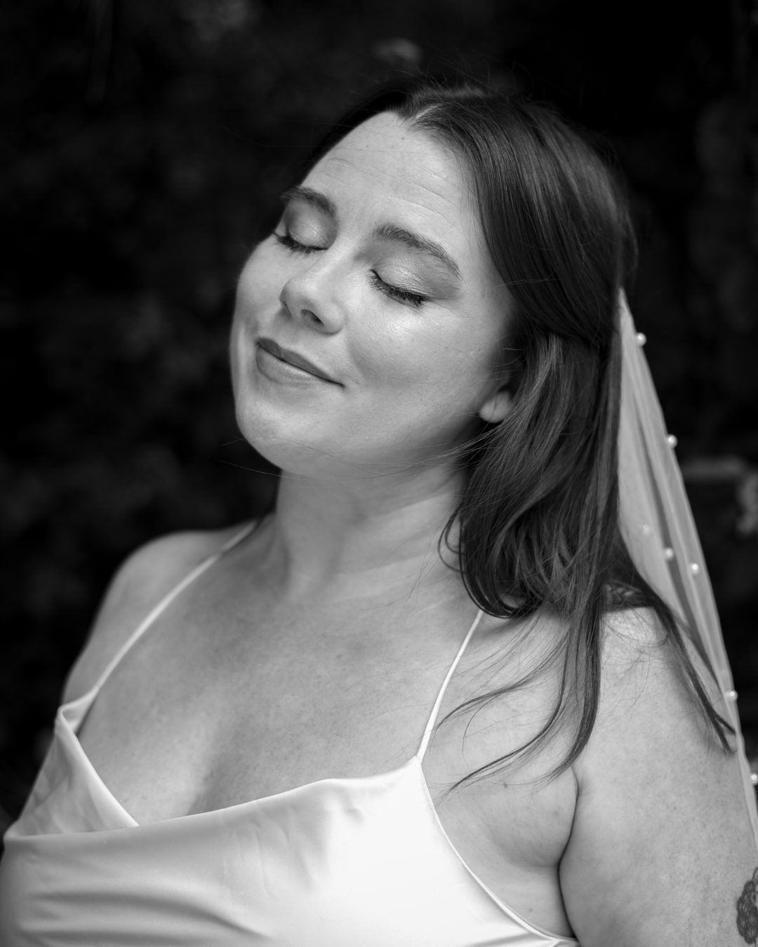 A woman with closed eyes and a slight smile, wearing a dress and a veil with pearl accents, with her hair down, outdoors.