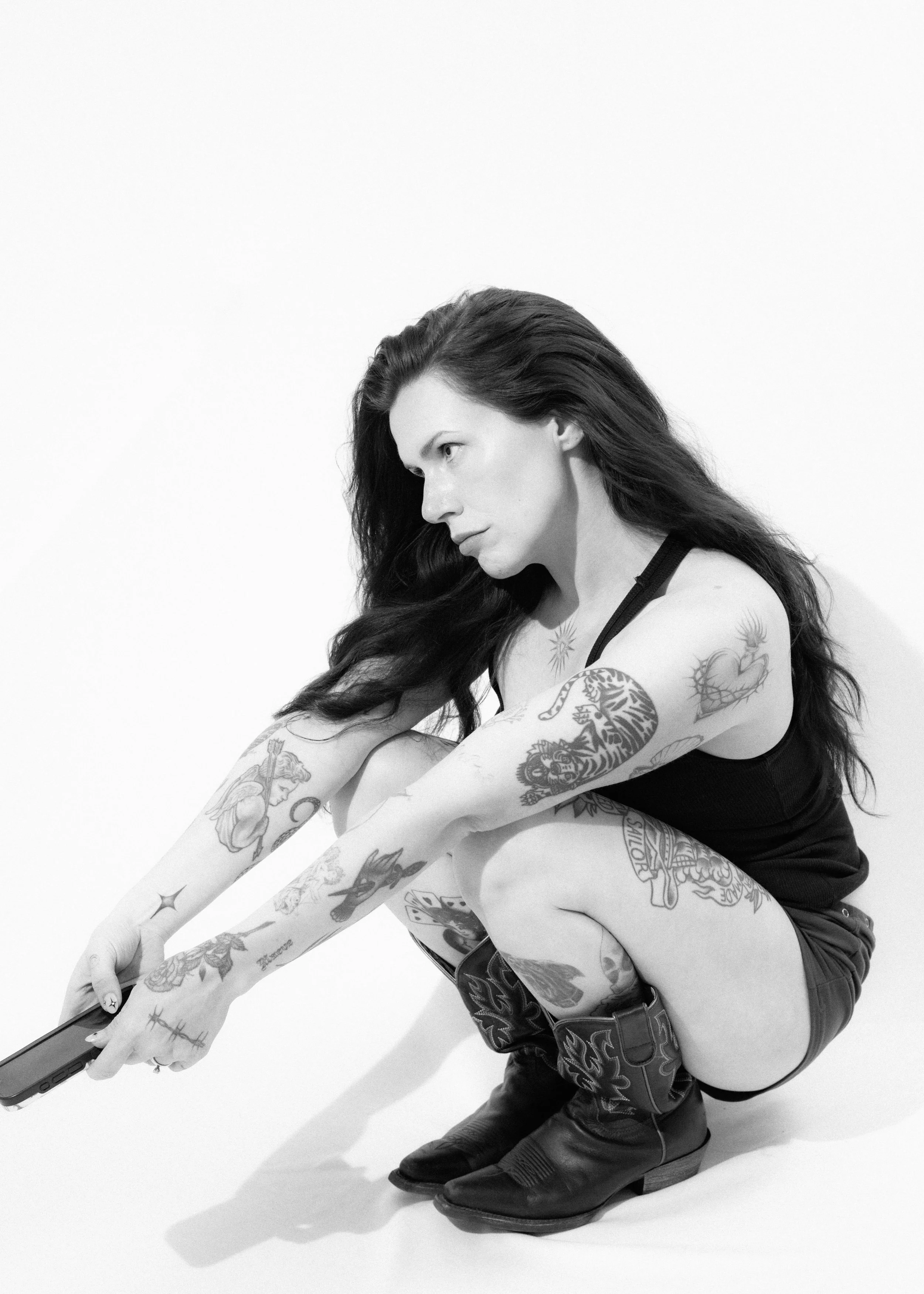 A woman crouching and stretching forward, holding a tablet device, with long dark hair, tattoos on her arms, wearing a black tank top, shorts, and black boots, against a plain white background.