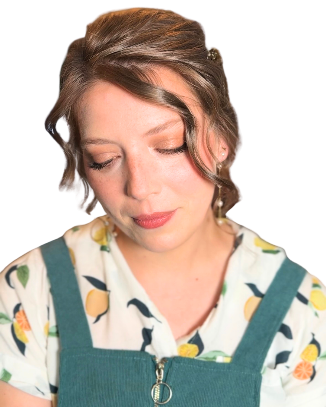 A woman with short, wavy brown hair and light makeup, wearing a white blouse with a lemon print and green overalls, looking down with a serene expression.