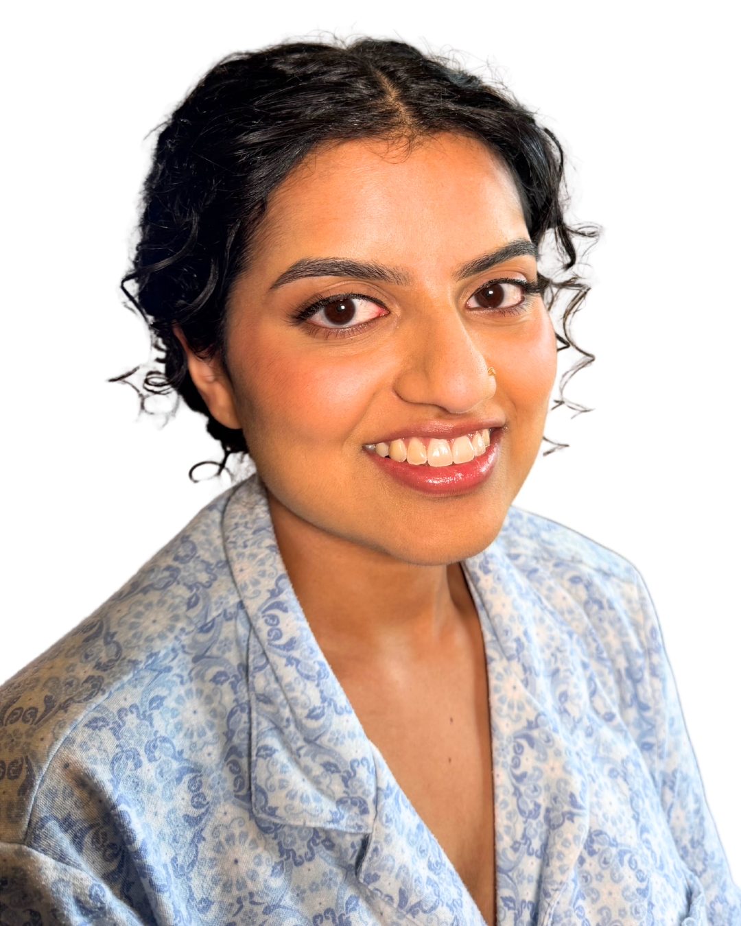 A woman with dark, curly hair, fair skin, and brown eyes smiling, wearing a light blue patterned shirt, against a black background.