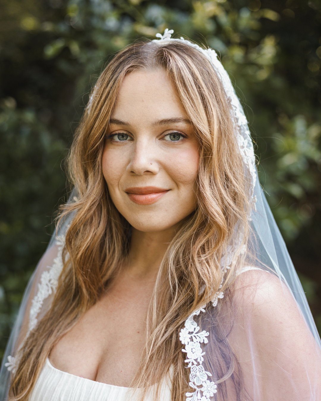 A woman with long, wavy, light brown hair and blue eyes wearing a white dress and lace veil, outdoors with green foliage in the background.