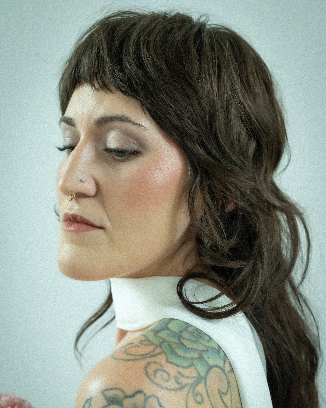 Side profile of a woman with dark brown hair, tattoos on her shoulder, and wearing a white sleeveless top.