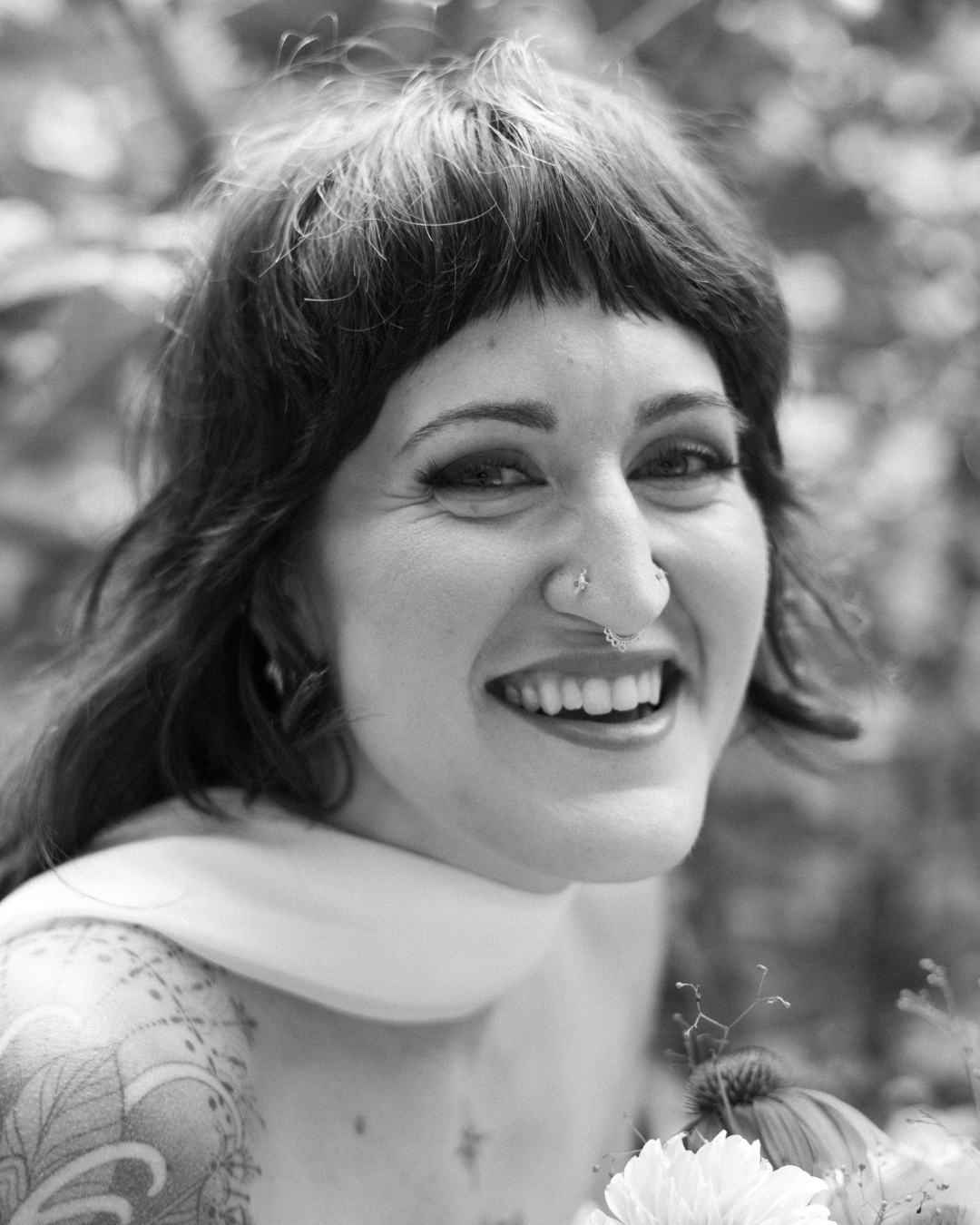 A smiling woman with short, dark hair, tattoos on her arm, and facial piercings, outdoors with a blurred background.