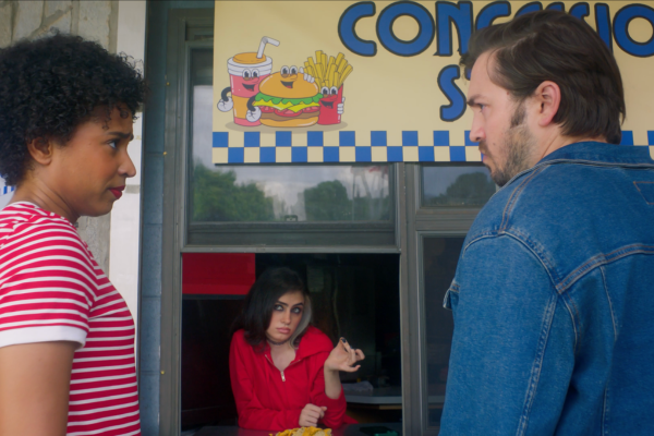 A woman in a red hoodie standing in a food stand window, making a skeptical expression, while two people, a woman with curly hair in a red and white striped shirt and a man with dark hair and a blue denim jacket, stand outside facing her. A colorful 