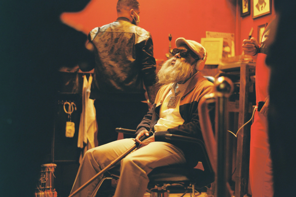 An elderly man with a long white beard, wearing a cap, glasses, a jacket, and tan pants, sitting in a wheelchair in a room with red walls, surrounded by two younger men and some decorative items on the wall.