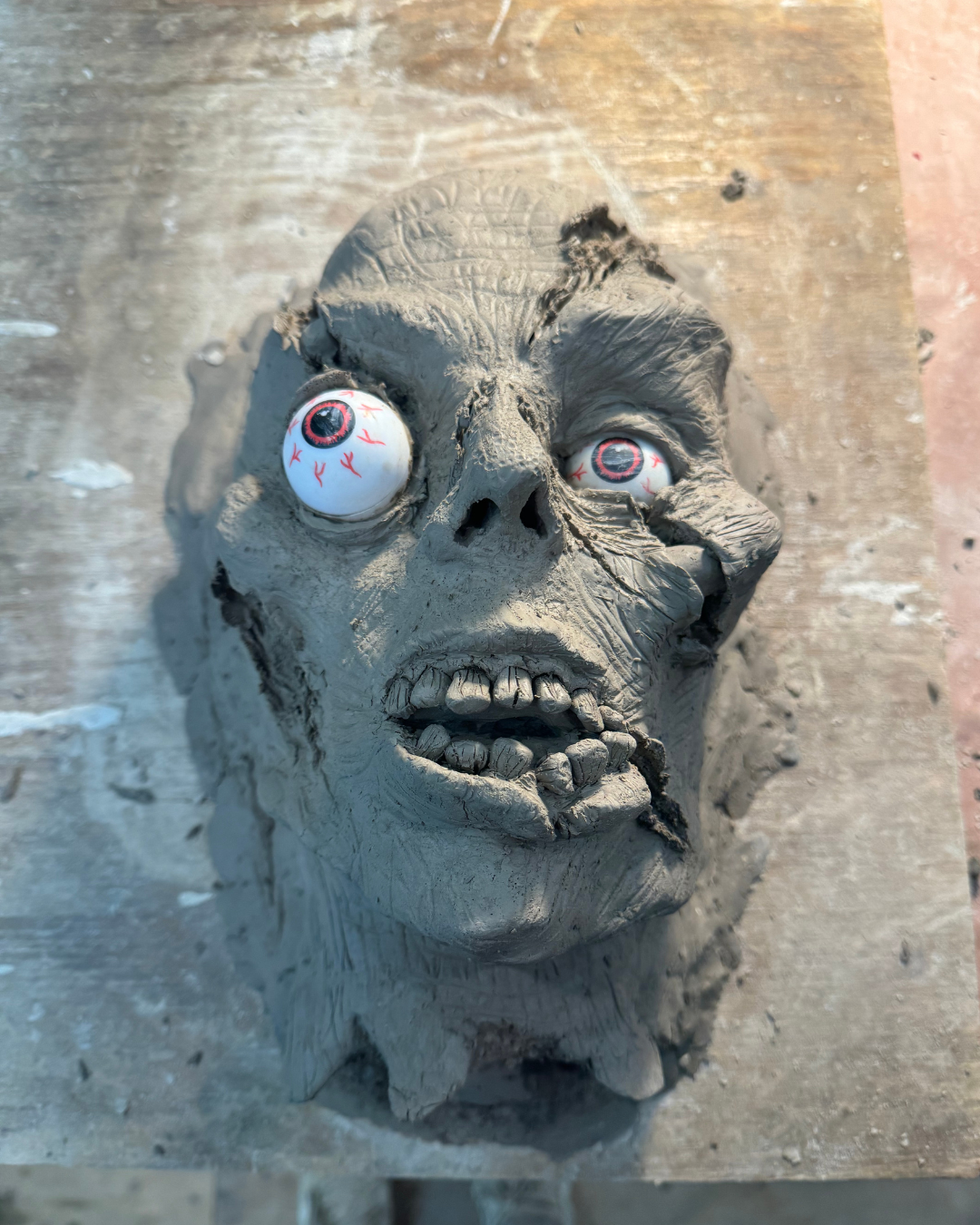 A clay sculpture of a zombie face with protruding eyeballs and open mouth, placed on a wooden surface.