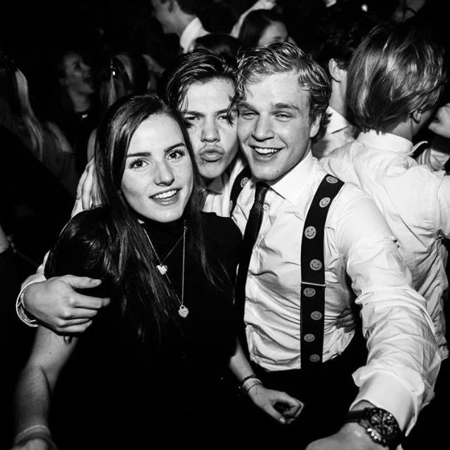 Drie jongeren poseren samen in een zwart-wit foto, omringd door andere mensen op een feest of evenement.