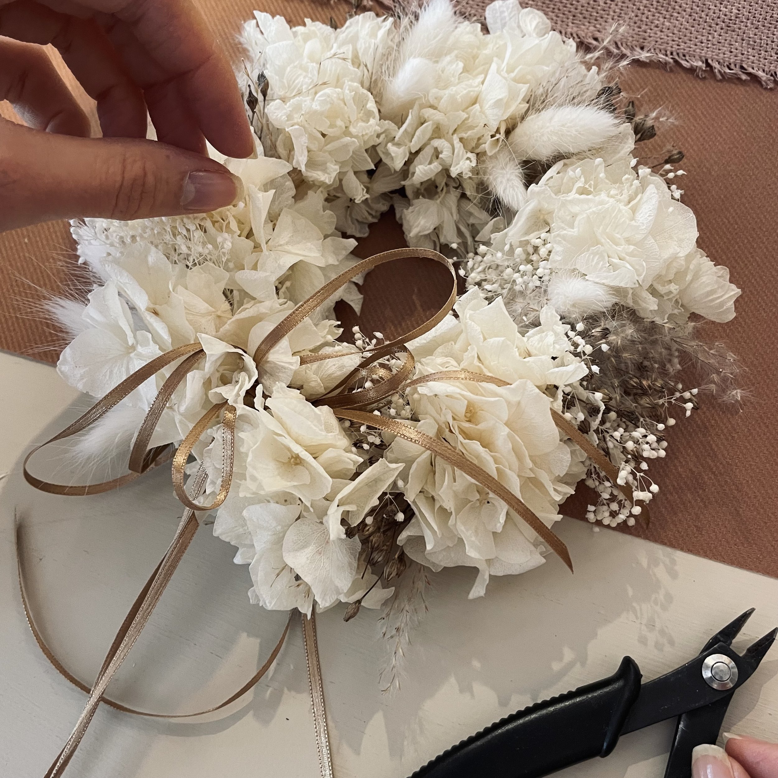 Weiße Blumenkranz mit braunen Bändern, auf einem Tisch, mit einer Hand, die ihn befestigt, und einer Zange rechts im Bild.