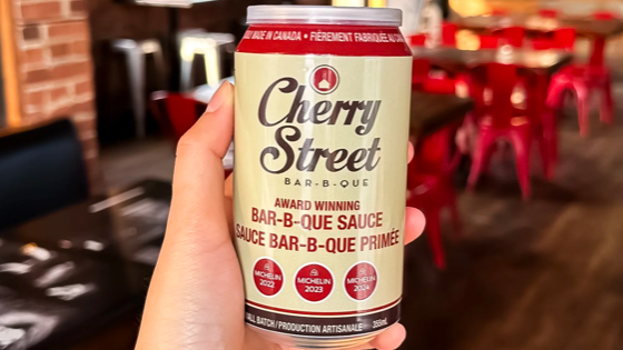 Person holding a Cherry Street Bar-B-Que sauce can in a restaurant setting with red chairs and tables in the background.