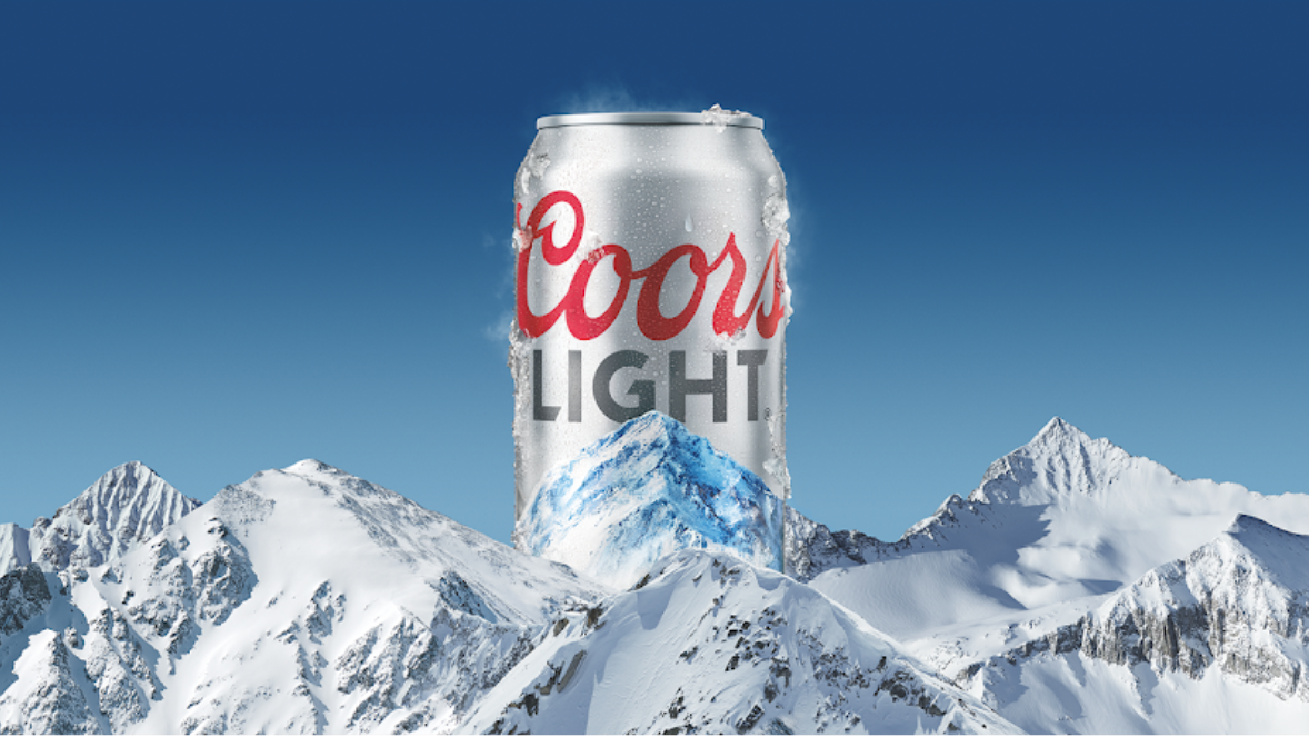 A large Coors Light beer can rises above snow-covered mountain peaks against a clear blue sky.