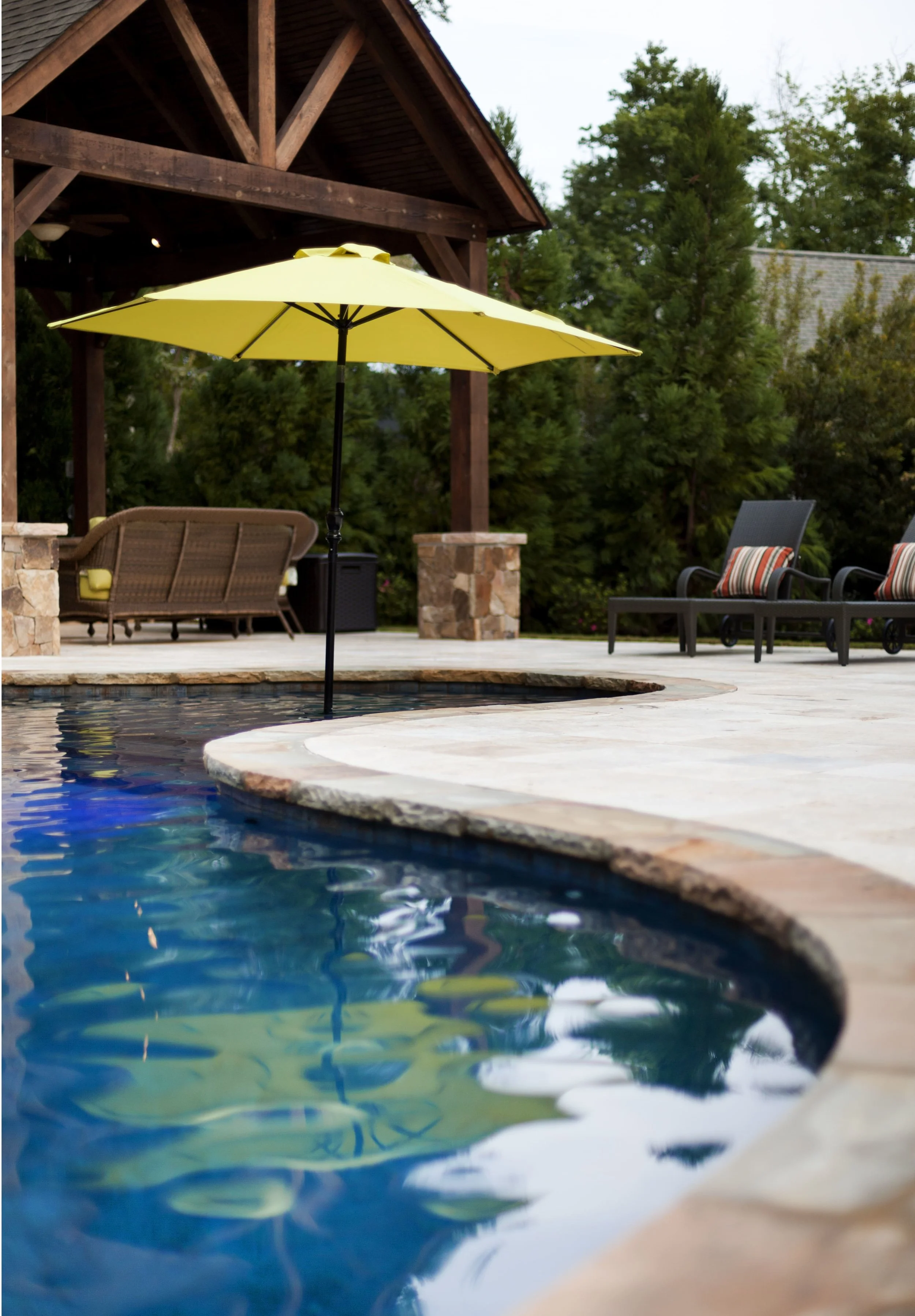 Chiseled edge on the stone coping ties in to the rustic feel of all the stained wood around. The umbrella that is in the pool provides shade for those who want it, while still allowing them to be in the pool.