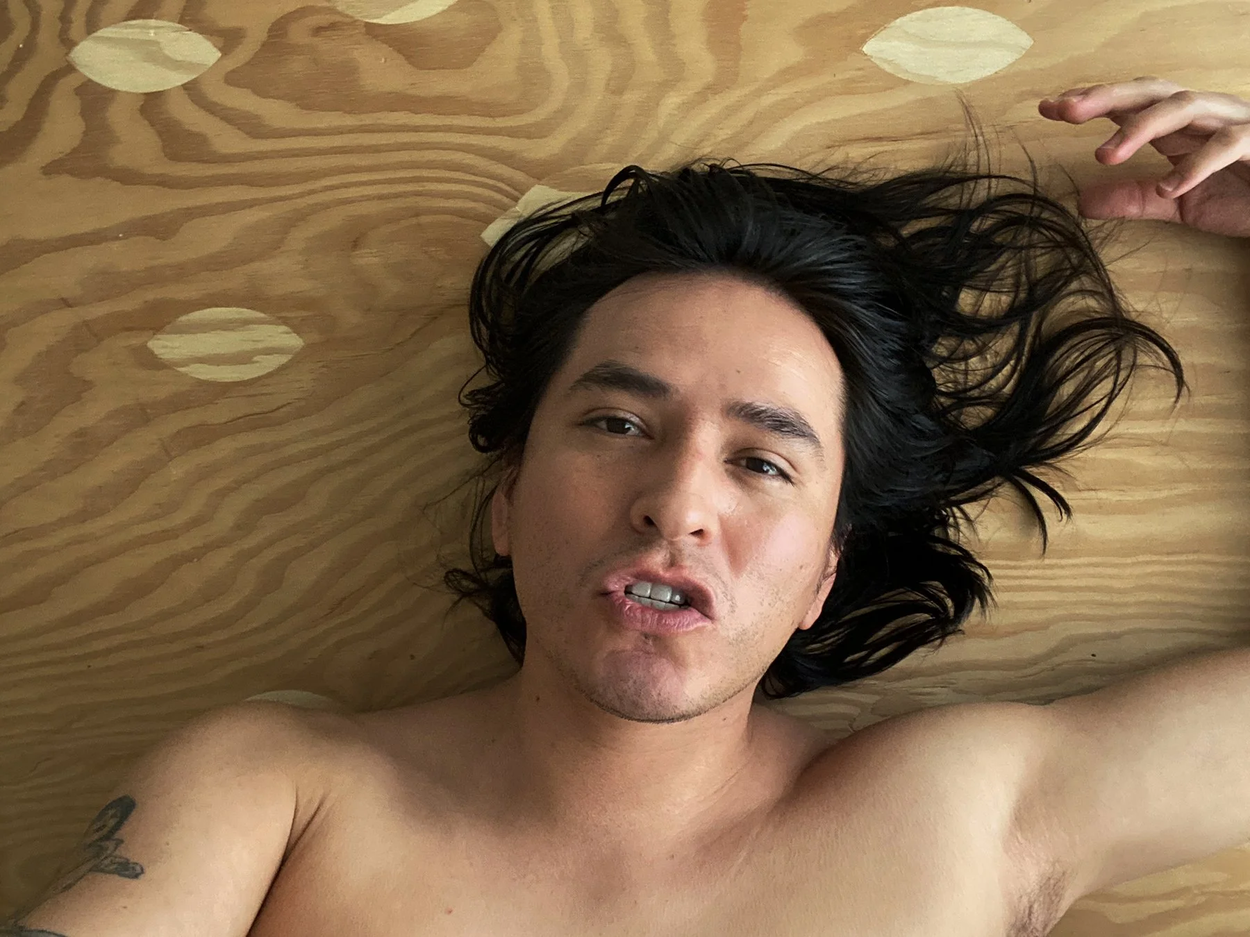 Demian DinéYazhi’, a Diné artist and activist, lying on a wooden floor with long black hair flowing around their head, gazing at the camera.