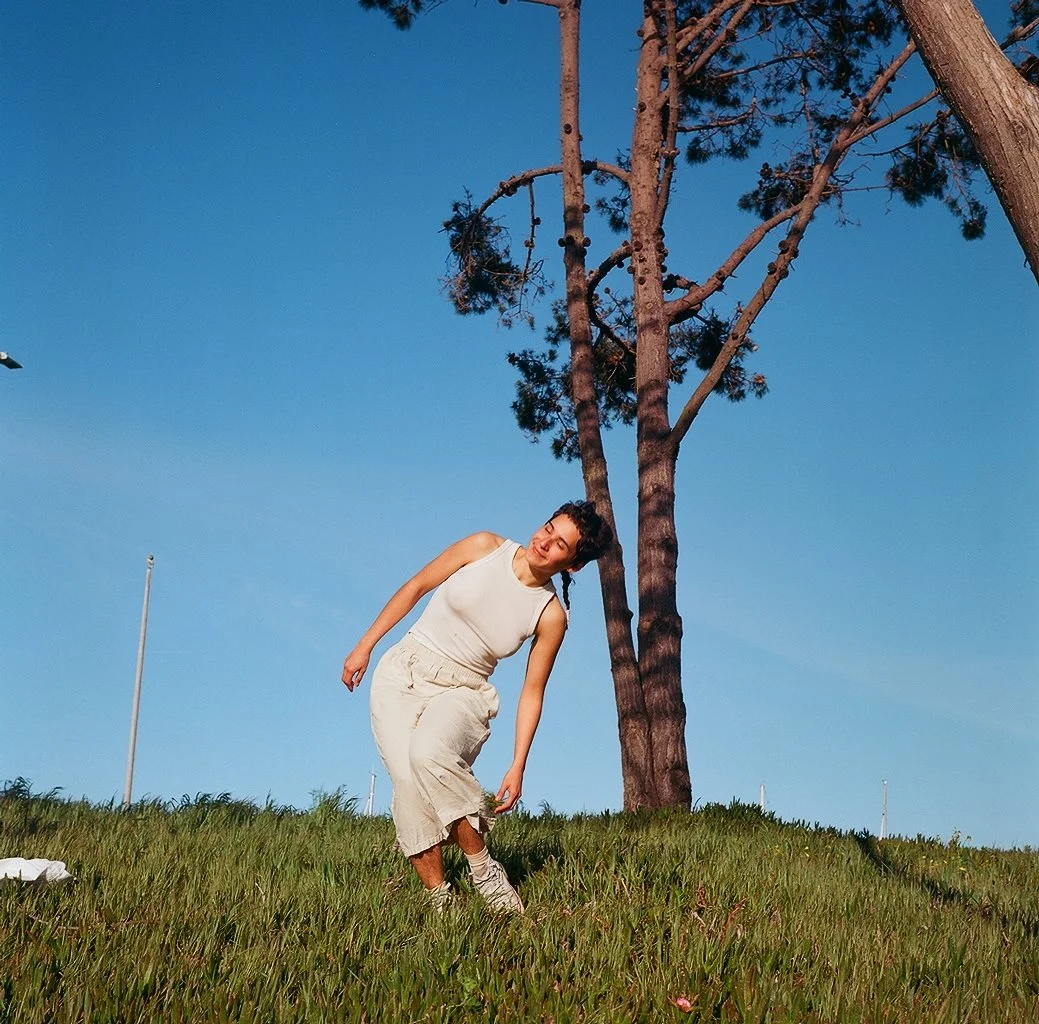 An indigiqueer artist named Yoli standing on a grassy hill, leaning slightly to the side, dressed in a light-colored outfit under a clear blue sky with trees in the background.