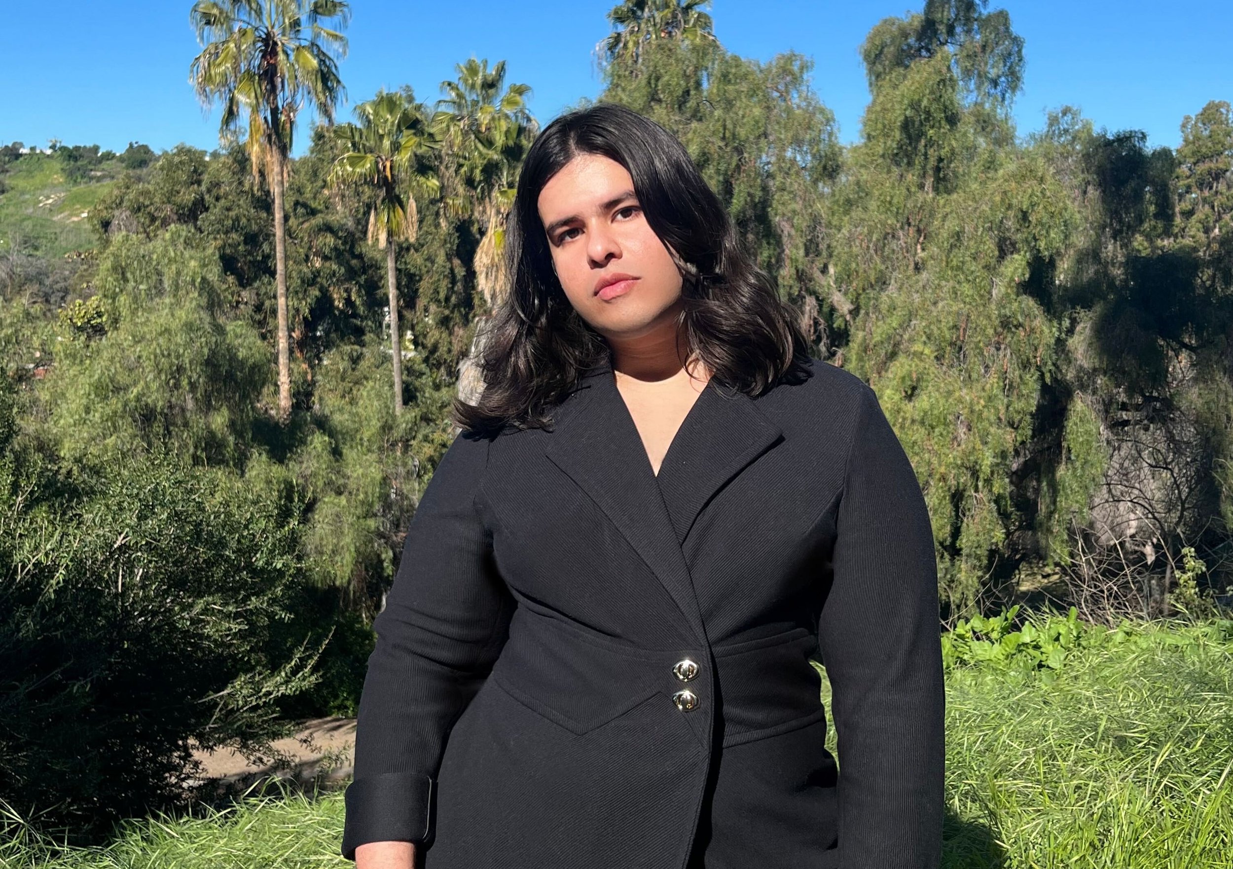 Portrait of Kira Xonorika, a femme artist, standing confidently in a natural setting with trees and palm trees in the background, wearing a black outfit.