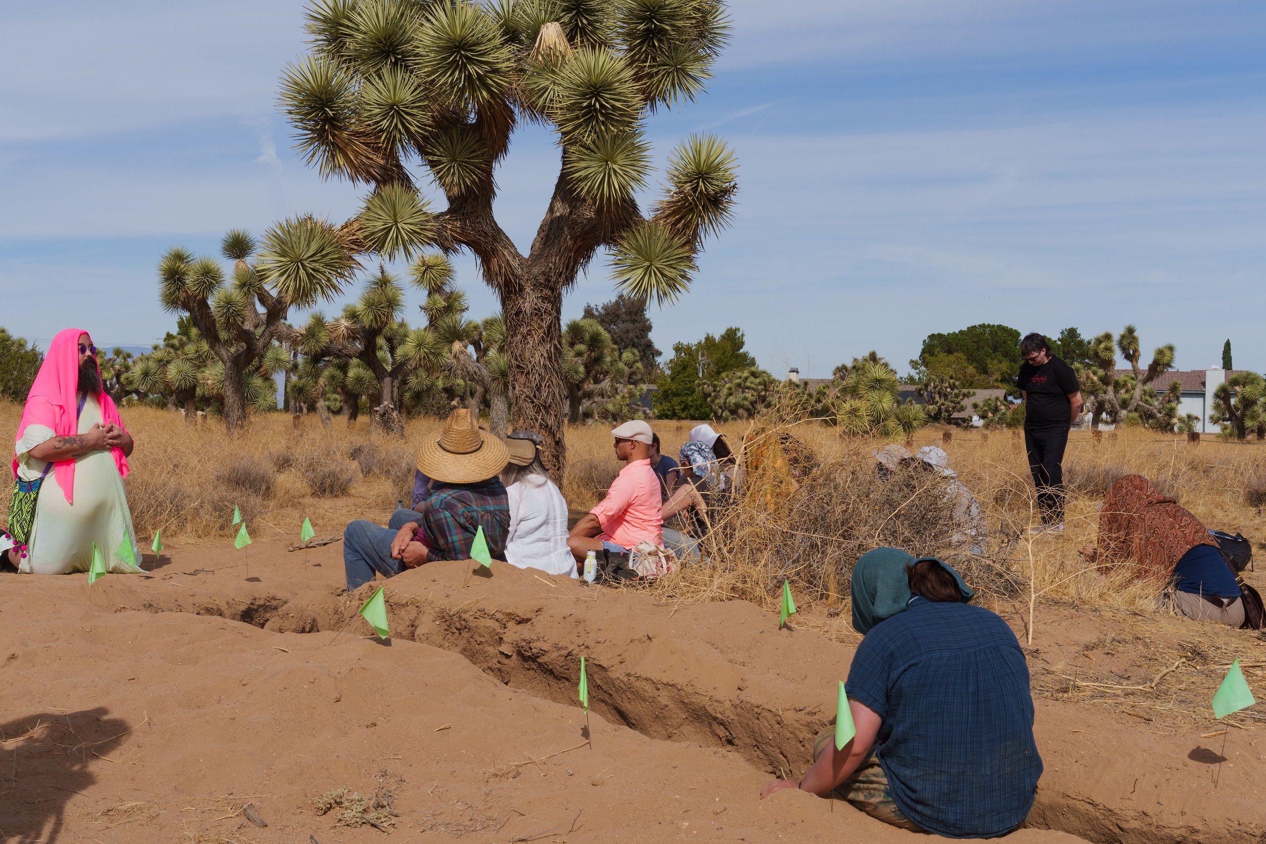 Edgar Fabián Frías, wearing a bright head covering, leading a ceremonial gathering in the Joshua Tree desert. Participants sit in contemplation around shallow, marked trenches beneath the shade of a large Joshua tree.
