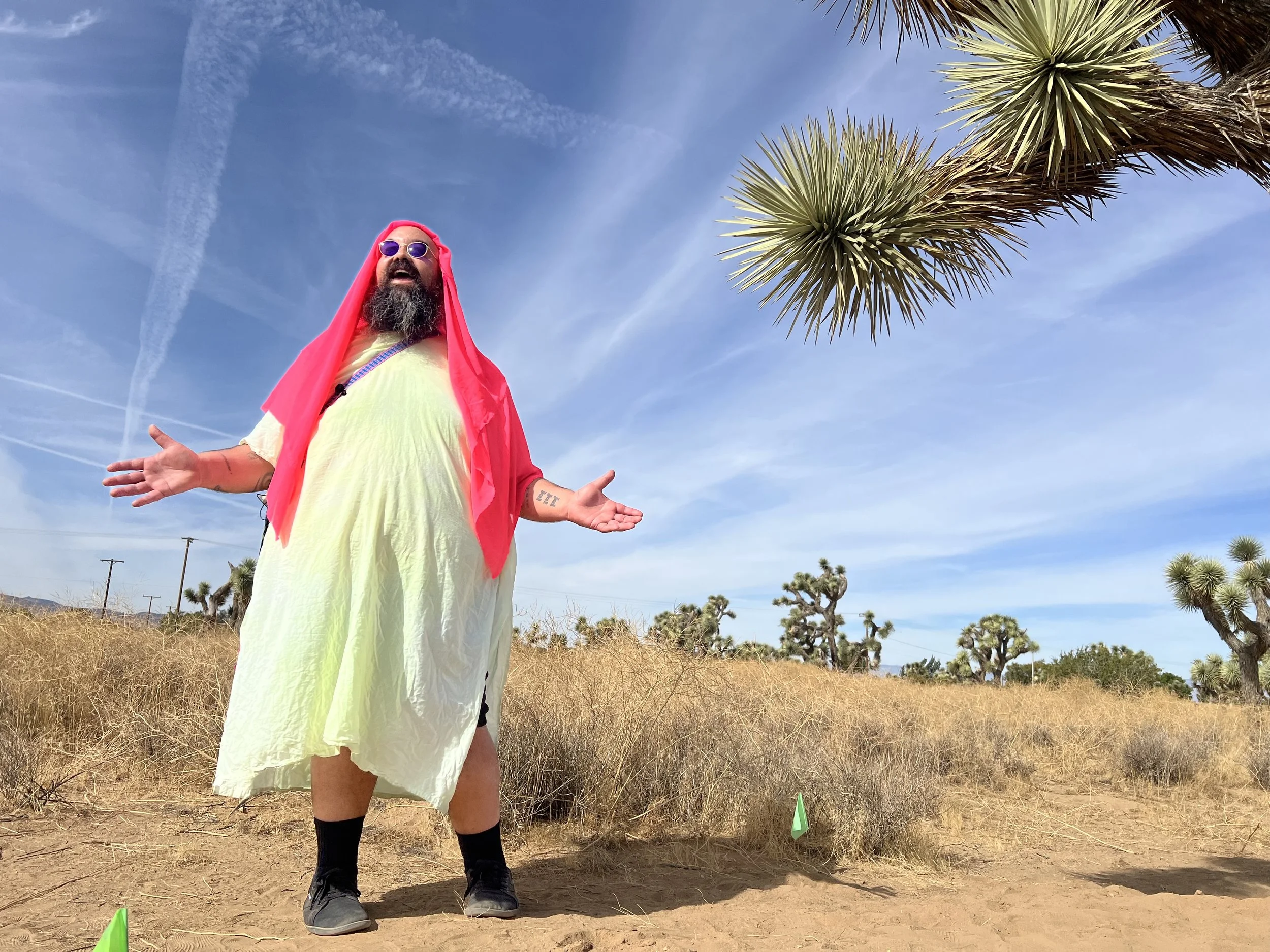 Person wearing a flowing, colorful garment and vibrant head covering, standing in an open Joshua Tree desert landscape with arms outstretched, looking upward. Clear blue sky and Joshua trees in the background.