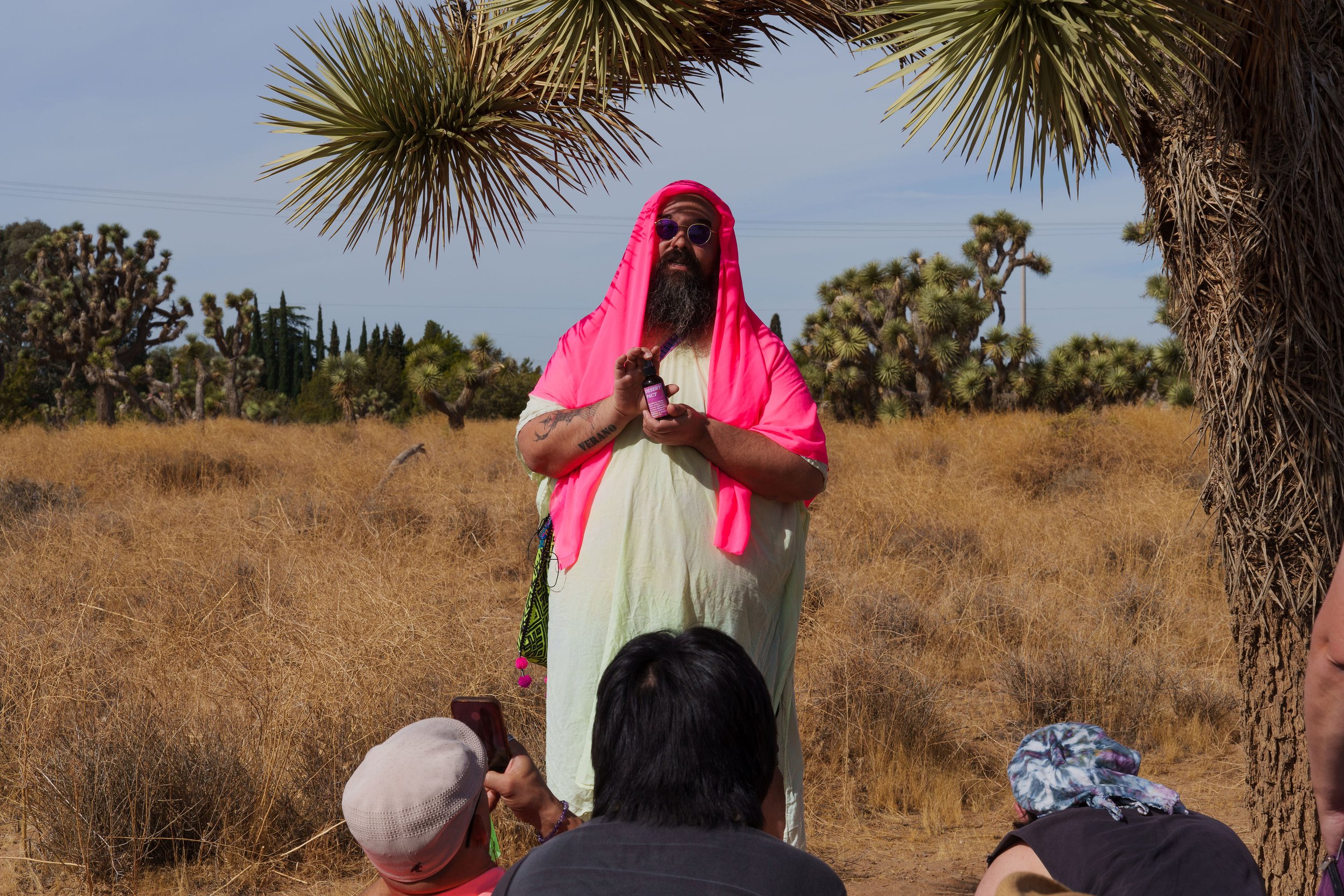 Edgar Fabián Frías, wearing a vibrant garment and head covering, leading a guided performance in the Joshua Tree desert, holding a small bottle as participants observe the ceremony closely.