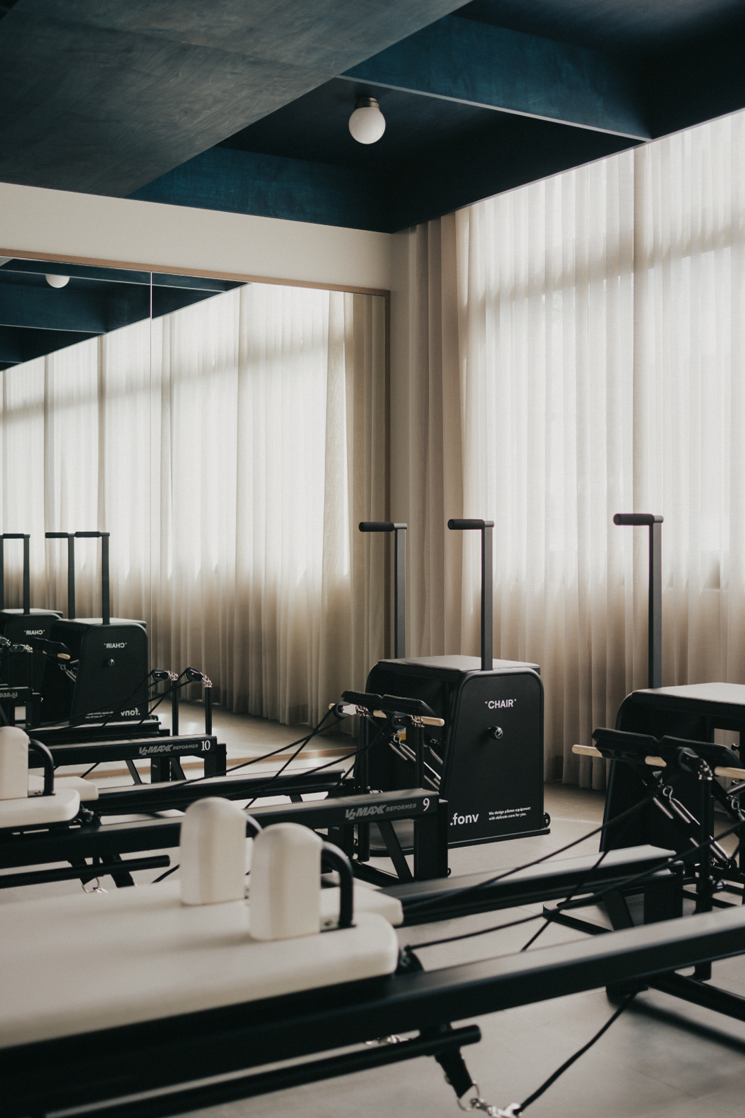 A Pilates or fitness studio with reformer machines near a large window with sheer curtains, and a ceiling with blue and gray panels.