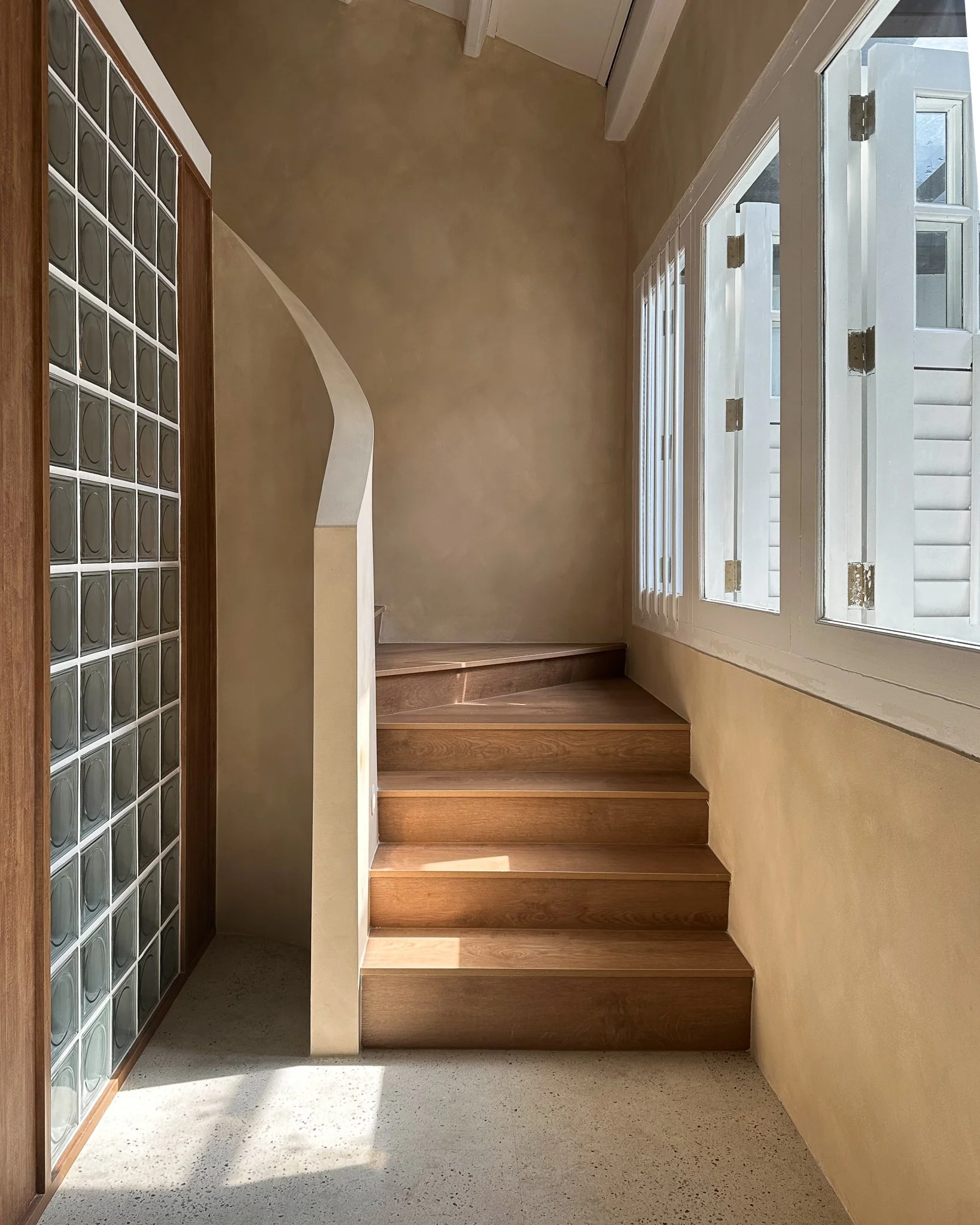 Interior of a house with wooden stairs leading up, a large window allowing natural light, and a glass block wall on the left.