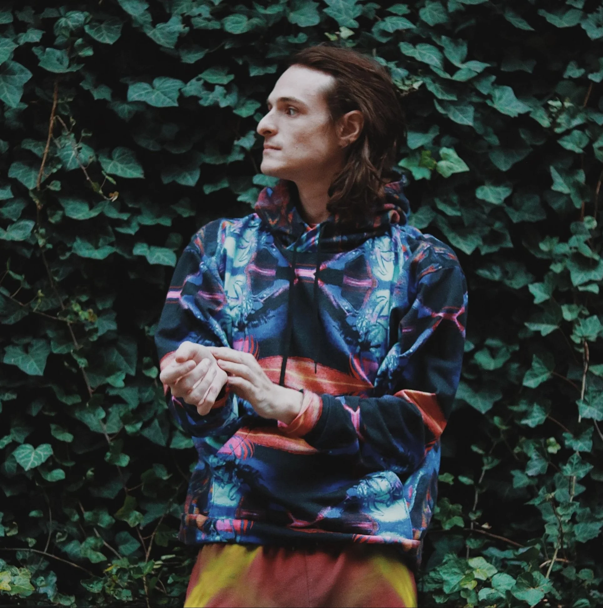 A person with long brown hair wearing a colorful hoodie standing in front of green leafy plants.