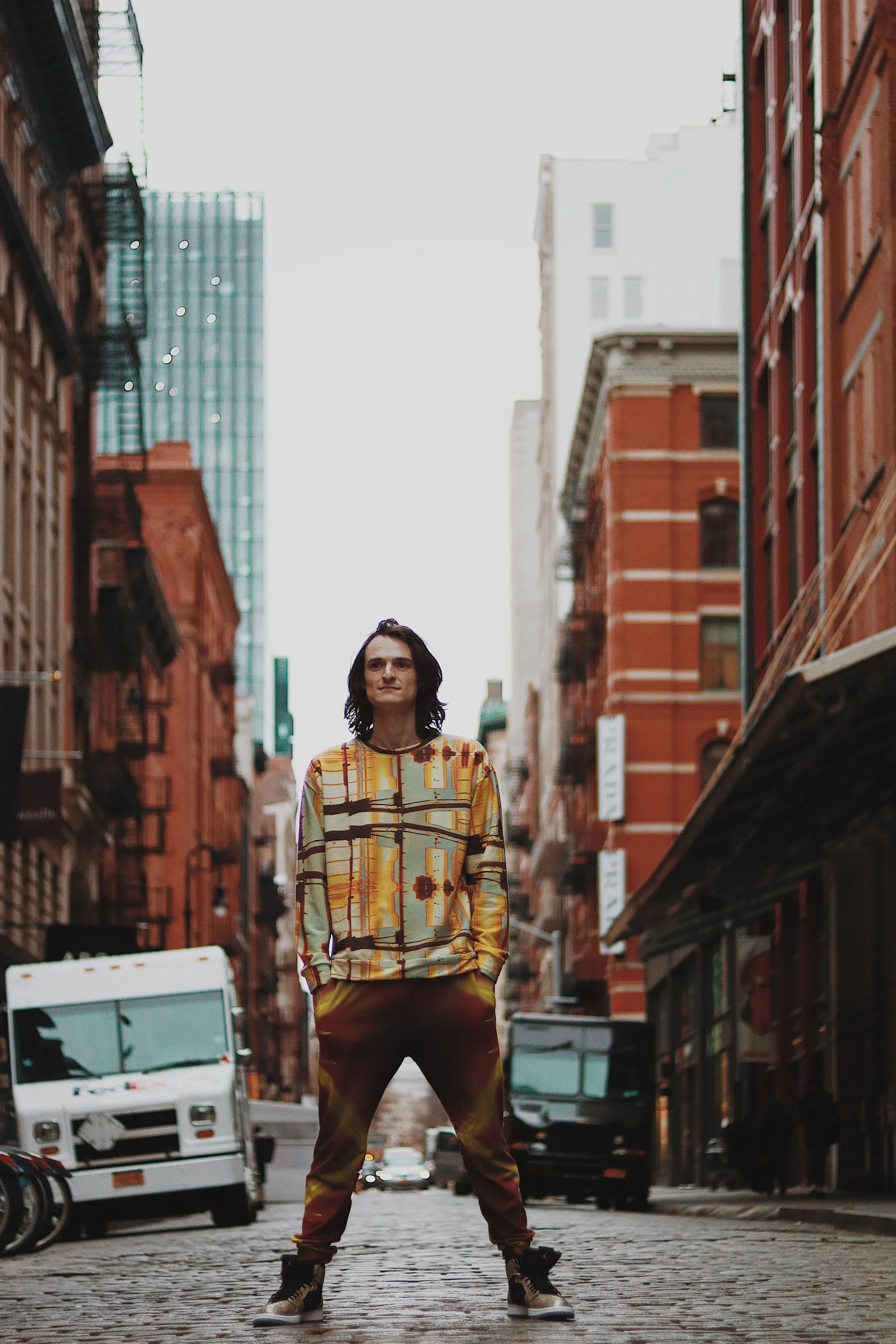 A person standing in the middle of a city street with tall buildings on either side, wearing a colorful patterned sweatshirt and pants, with hands in pockets and looking slightly to the side.
