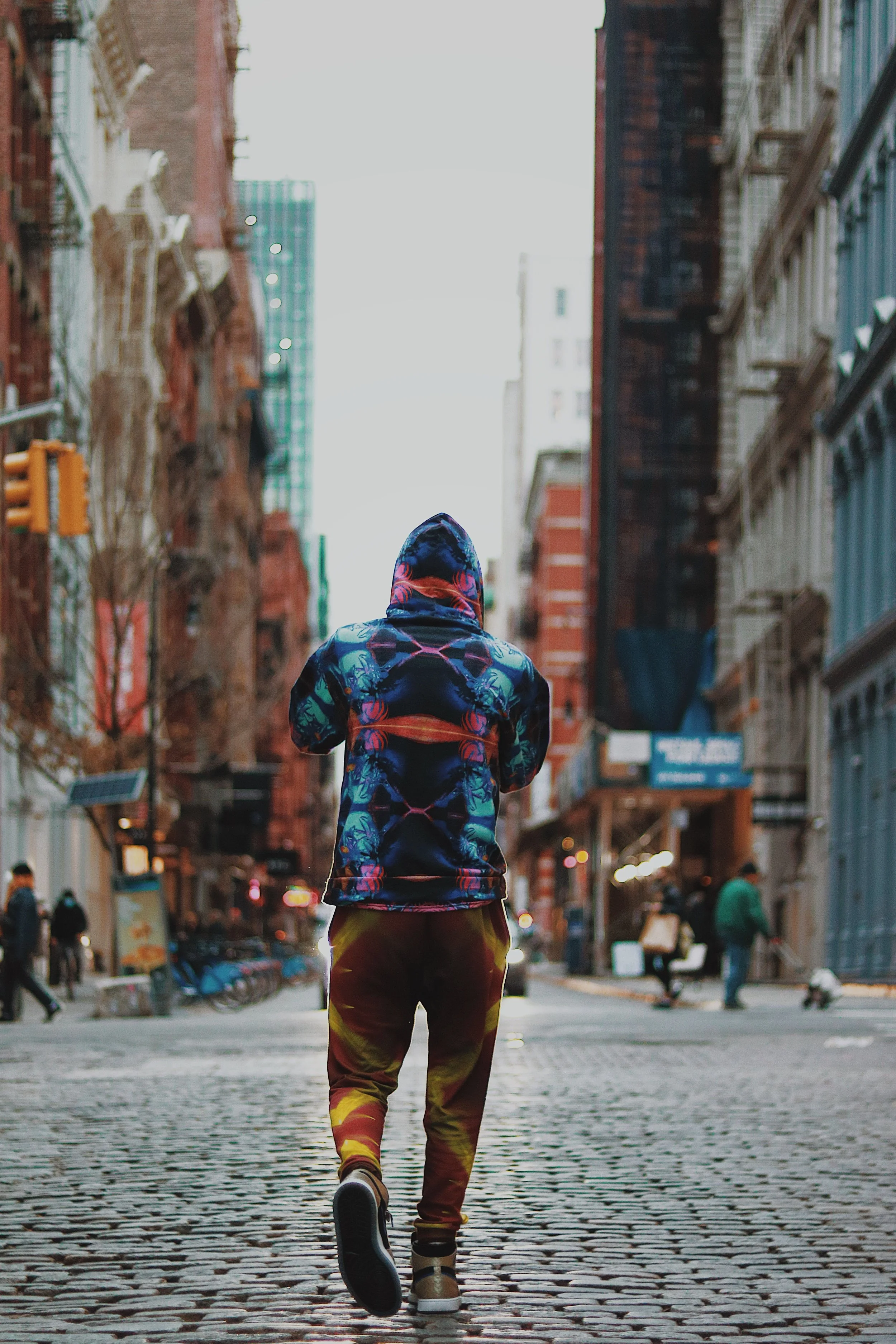 Person wearing a colorful hoodie walking on a cobblestone street in a city with tall buildings.