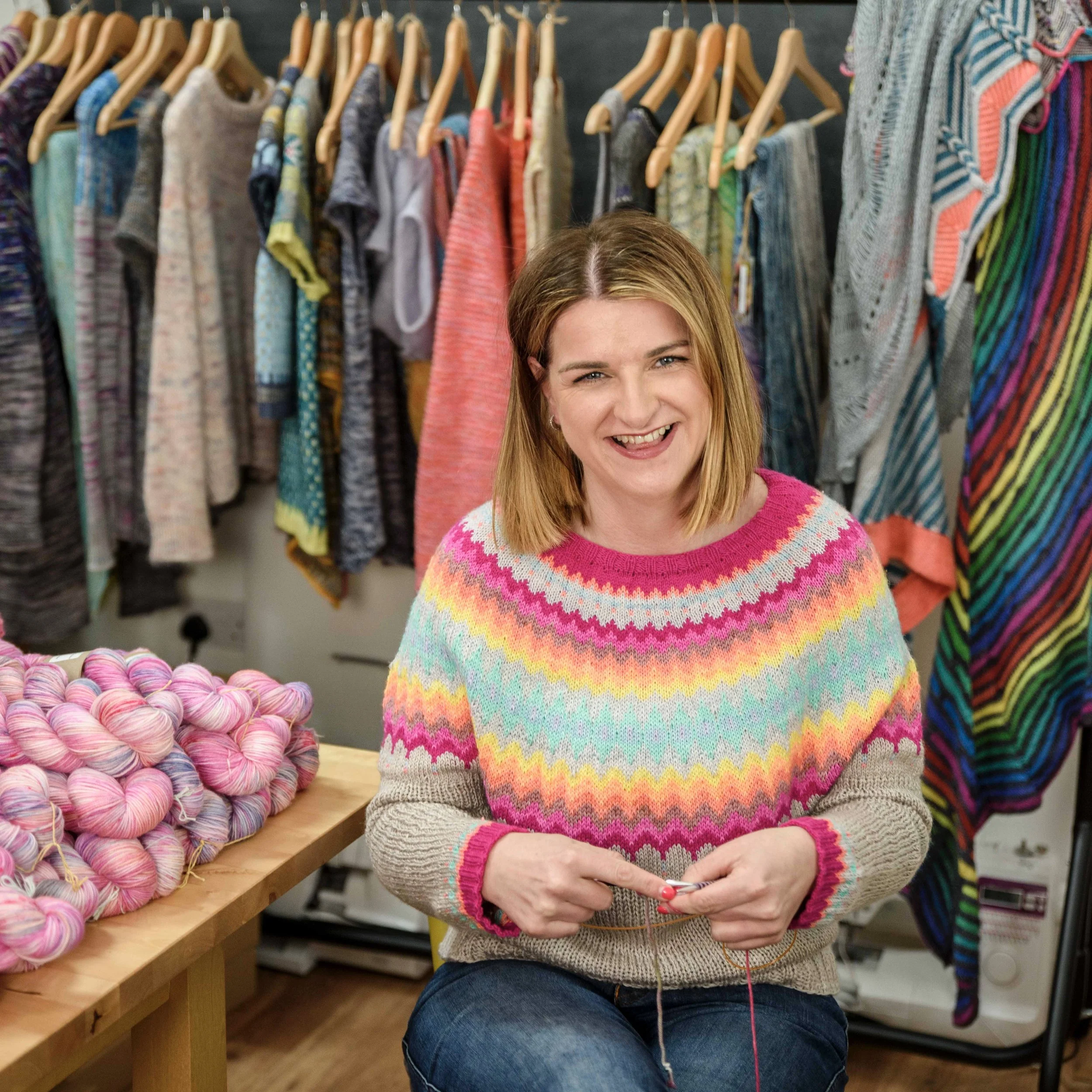 Claire sits on a chair with her knitting in her hands wearing a brightly coloured hand knit sweater