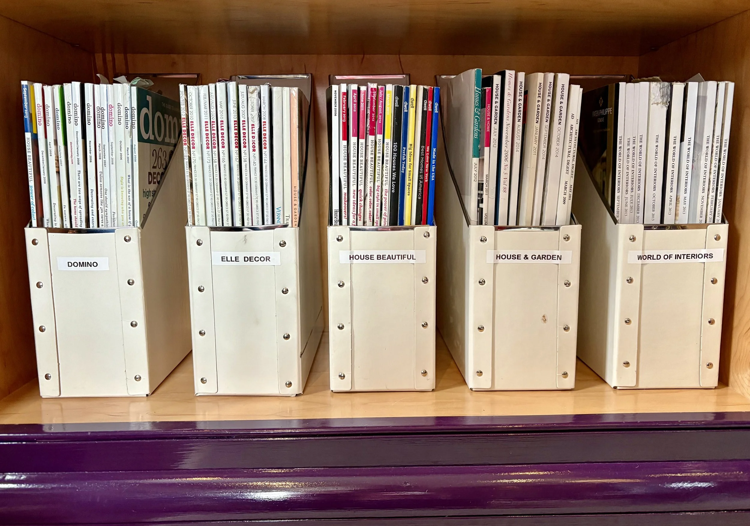 Books organized in white magazine racks labeled DOMINO, ELLE DECOR, HOUSE BEAUTIFUL, HOUSE & GARDEN, and WORLD OF INTERIORS on a wooden shelf.