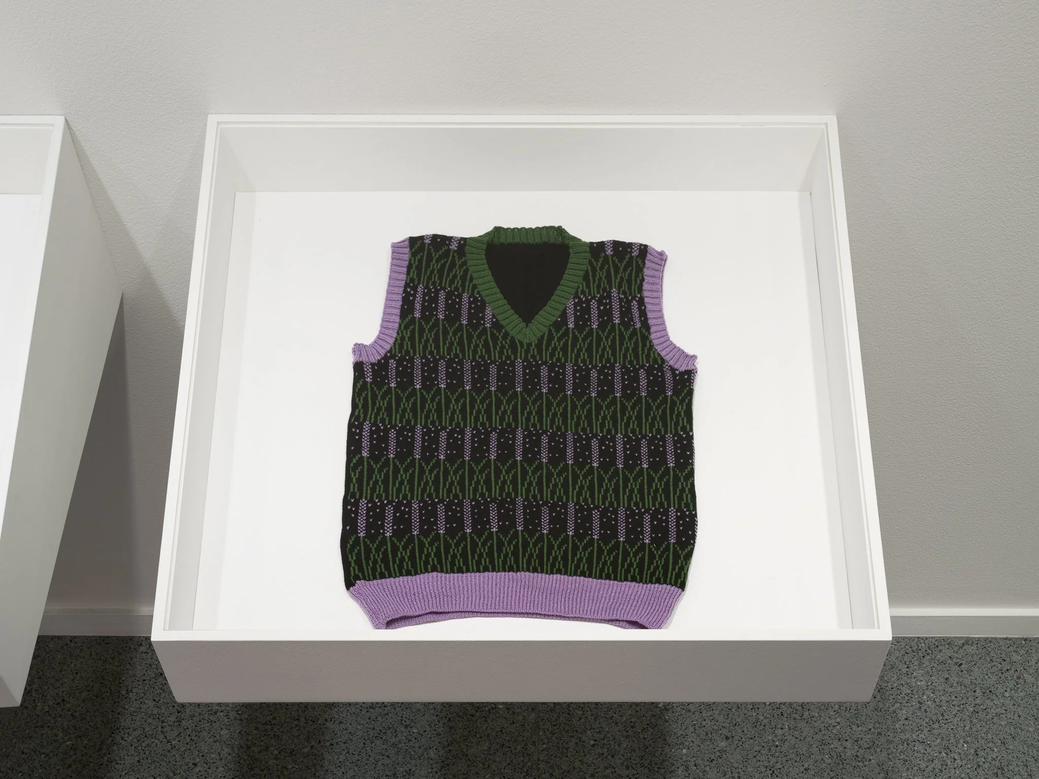 a dark sweater with green and purple patterns in a plexi box