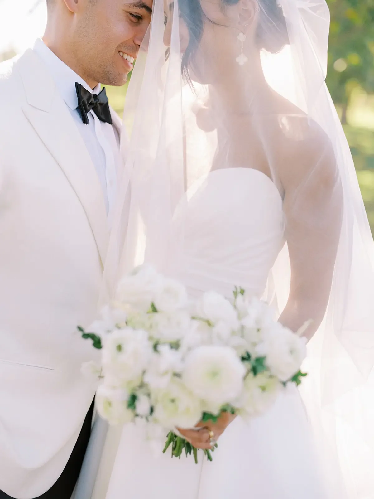 A newlywed couple stands close together outdoors, smiling with their foreheads lightly touching. The groom is dressed in a white tuxedo with a black bow tie, and the bride is wearing a strapless white wedding gown with a long veil covering her face. The bride holds a bouquet of white flowers, and the background is softly lit with greenery.