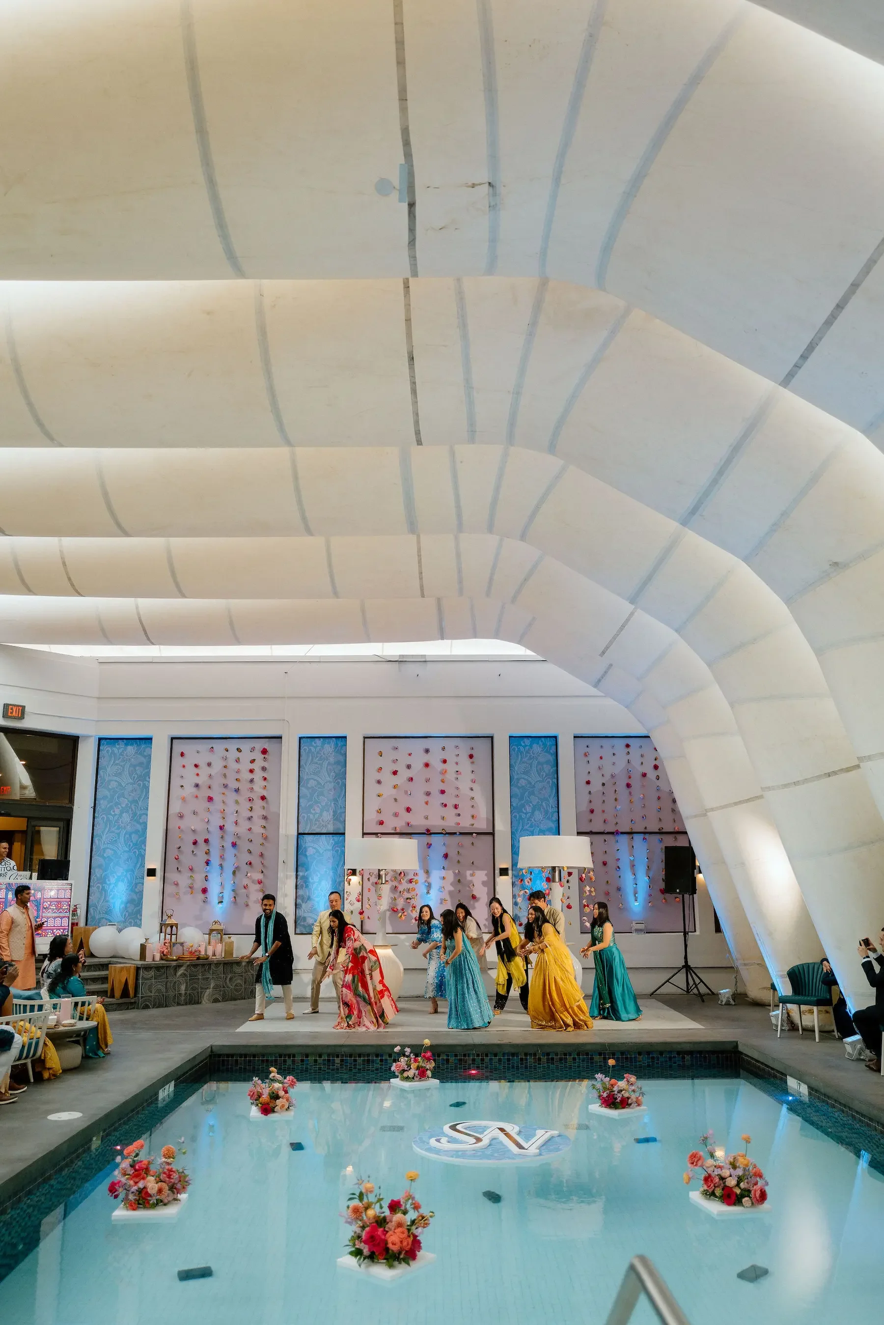 Indoor pool area decorated for celebration with a group of women dancing in colorful dresses, floral arrangements on the pool, and modern architectural ceiling design.