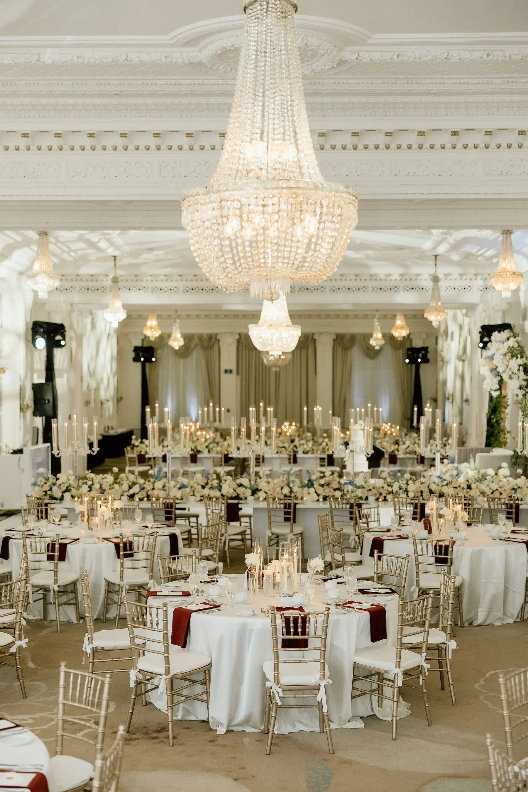 Elegant banquet hall decorated with large chandeliers, white tables with candles, floral centerpieces, and gold chairs for an event or wedding reception.
