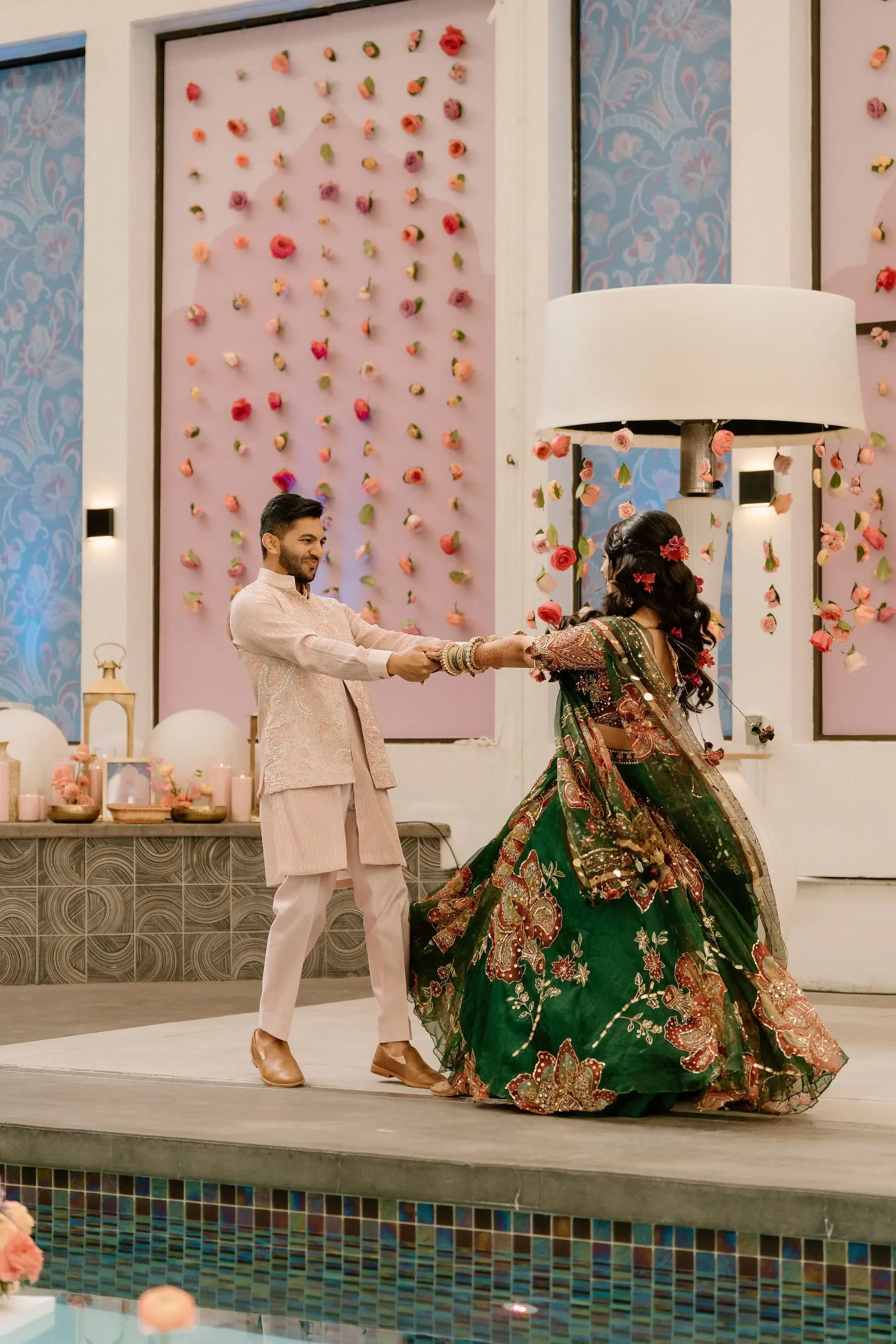 A man and woman dancing at a wedding celebration, with the woman wearing a traditional green embroidered outfit and the man in a light-colored sherwani, in a decorated hall with pink floral wall backdrop and candles.
