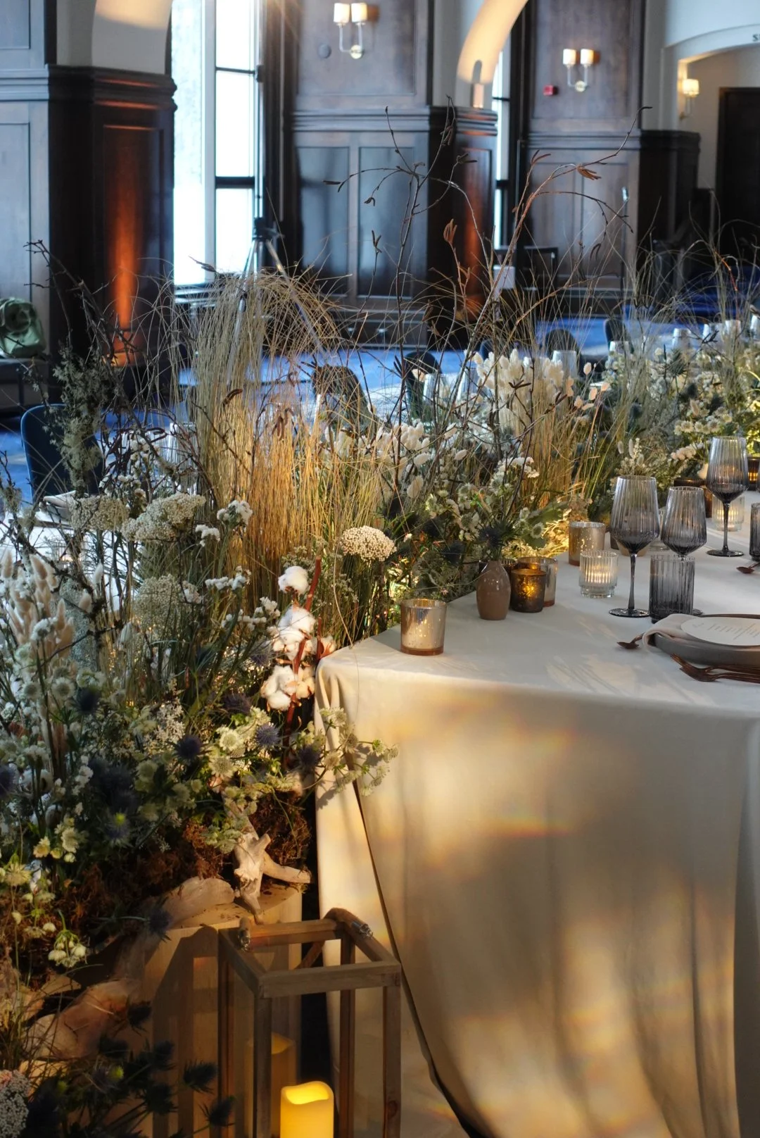 Elegant dining setup with a long table decorated with white and neutral-toned floral arrangements, candles, and dark glassware in a wood-paneled room with large windows and soft lighting.