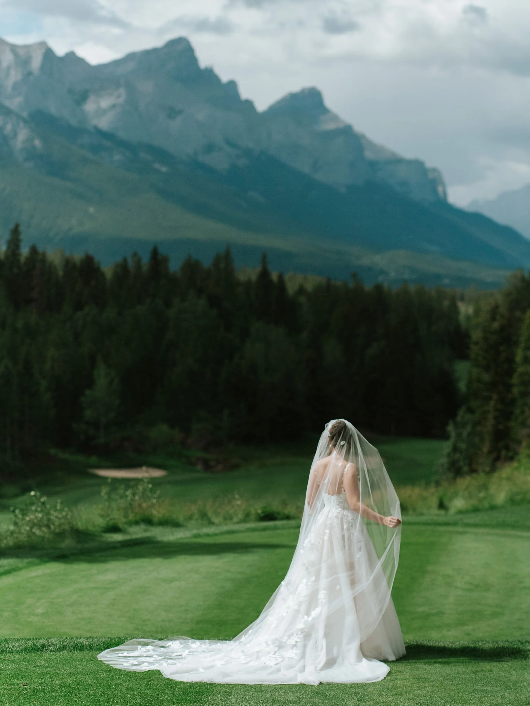 A bride in a white wedding dress and veil standing on a lush green golf course with a mountain range in the background.