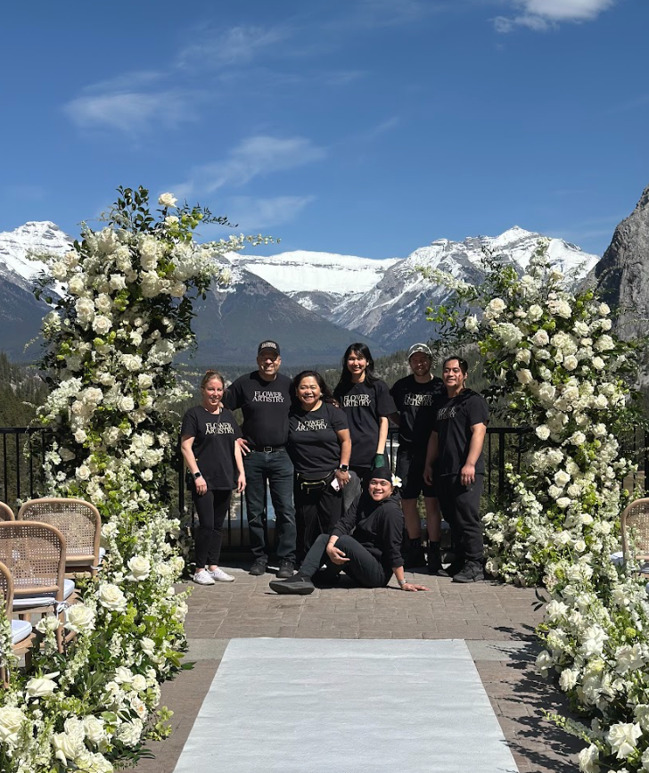 Group of seven people in black shirts with white text, standing and sitting in front of a floral wedding arch with snowy mountains and blue sky in the background.
