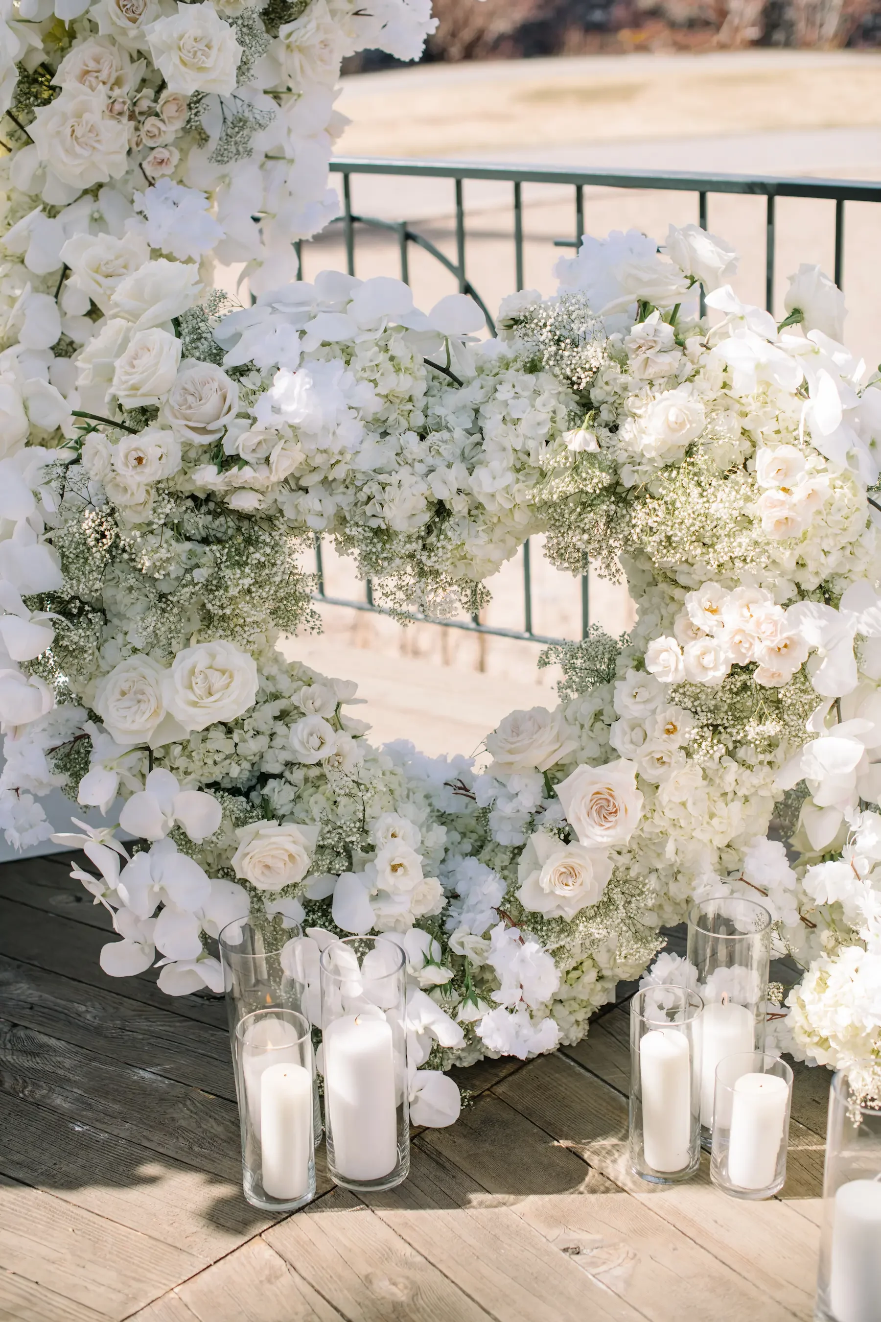 A white heart-shaped floral wreath made of roses, orchids, and baby's breath, surrounded by cylindrical glass candle holders with white candles on a wooden deck, outdoors.