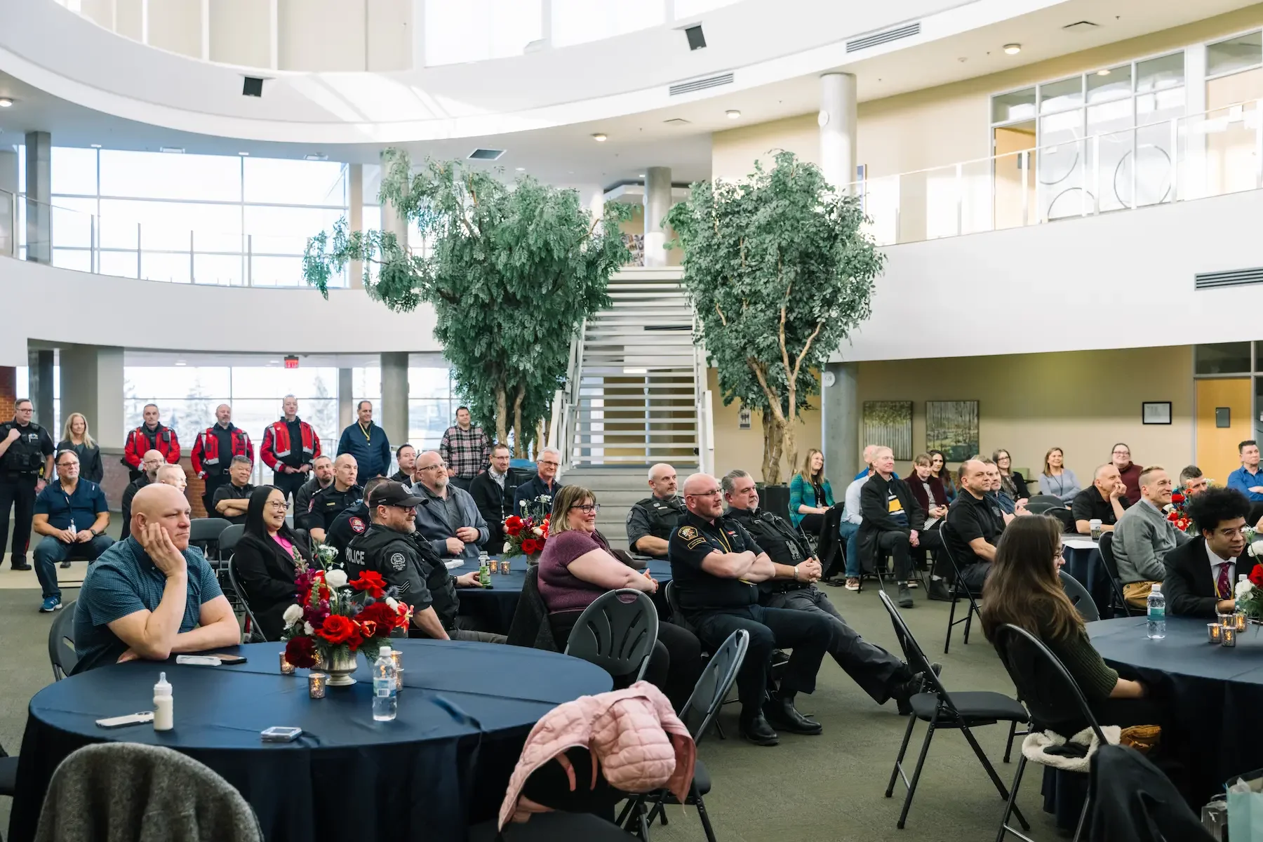Audience of police officers and civilians seated at round tables in a conference room with large windows, green trees, and indoor plants.