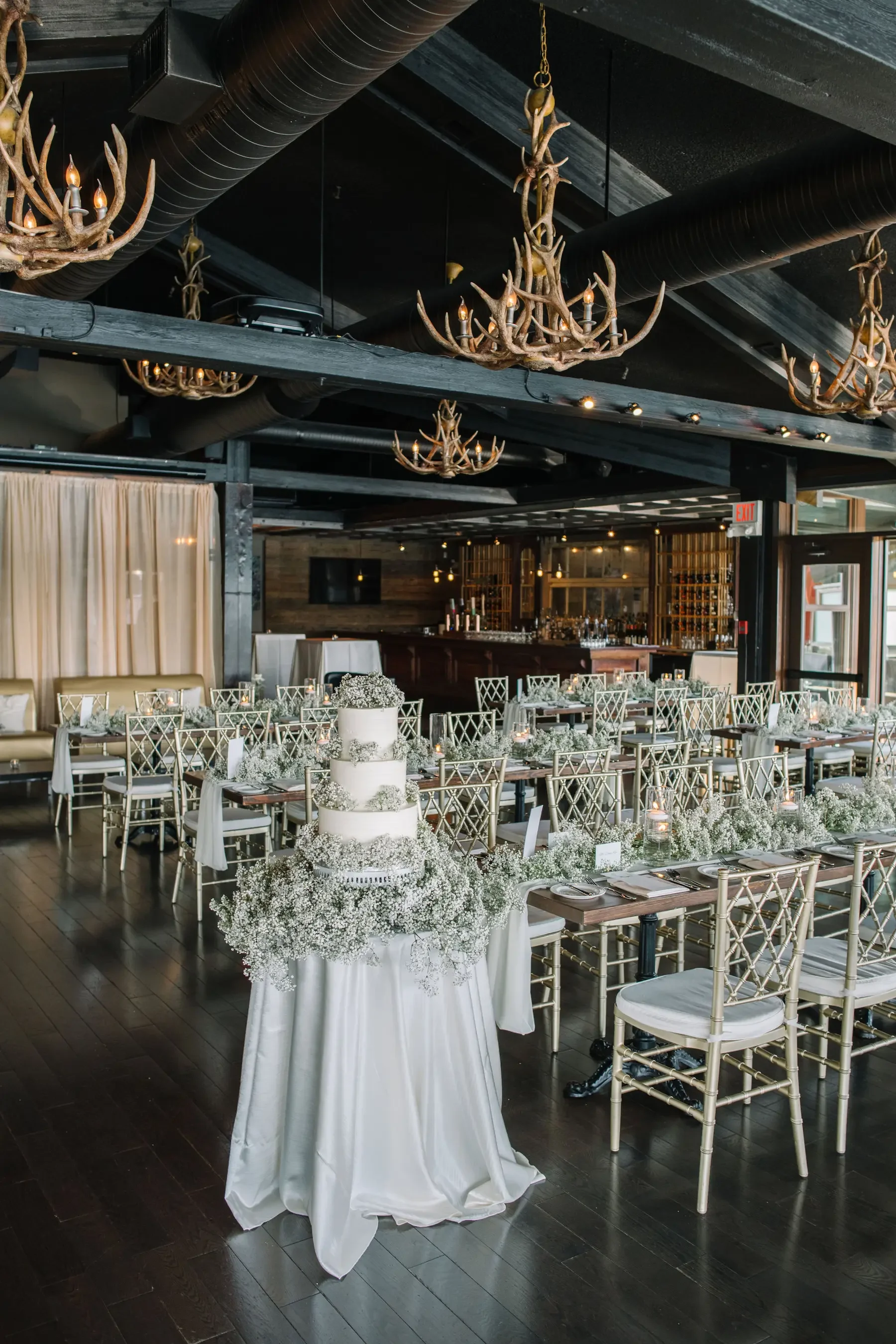 Wedding reception setup featuring a white tiered cake with baby's breath flowers on a table covered with white satin fabric, surrounded by elegant tables and gold chairs, with antler chandeliers hanging from the ceiling in a rustic style.