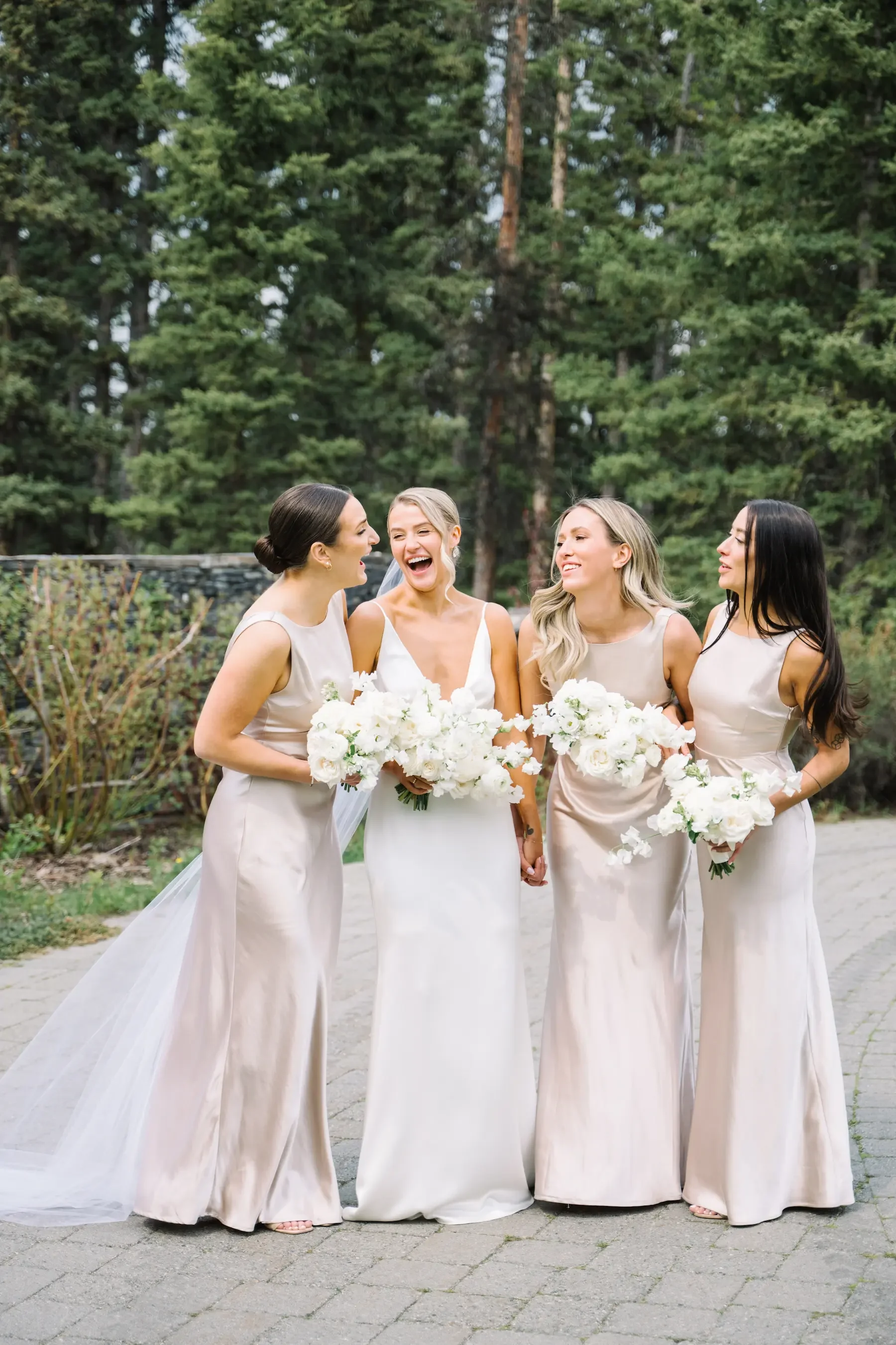 A bride and three bridesmaids standing outdoors, smiling and holding bouquets of white flowers, surrounded by green trees.