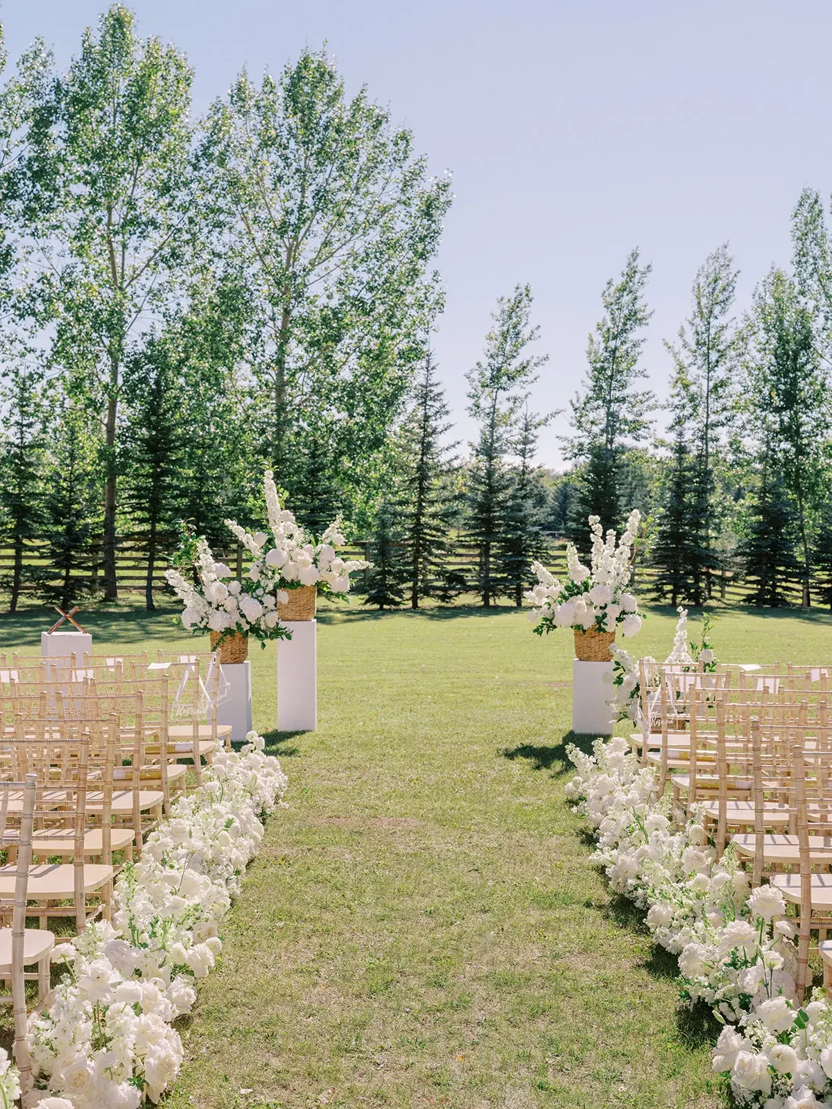 Outdoor wedding aisle decorated with white flowers and large floral arrangements in woven baskets on white pedestals, with trees in the background.