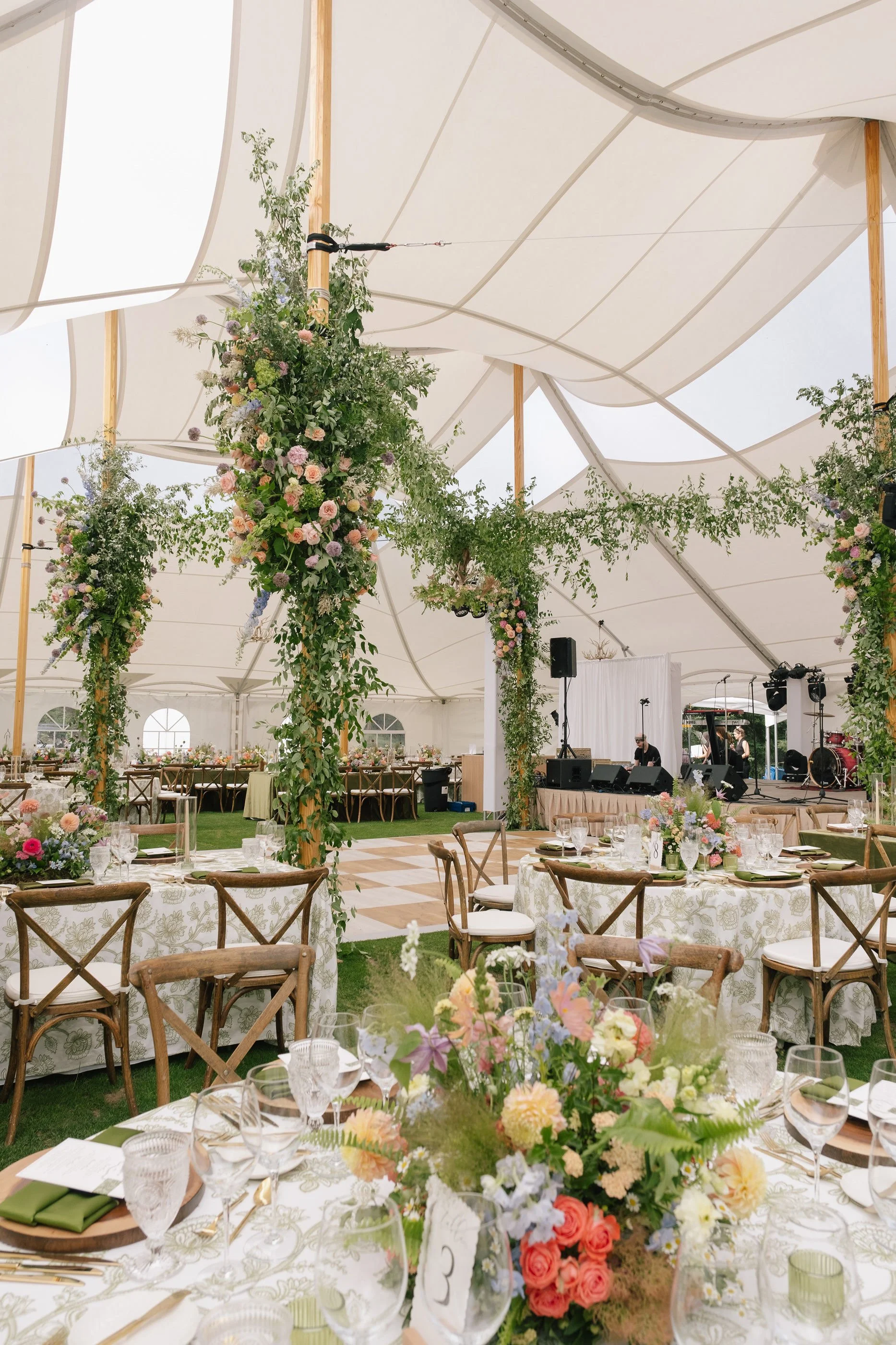 Inside a decorated wedding reception tent with floral arrangements, round tables with white tablecloths, and a stage with musical equipment.