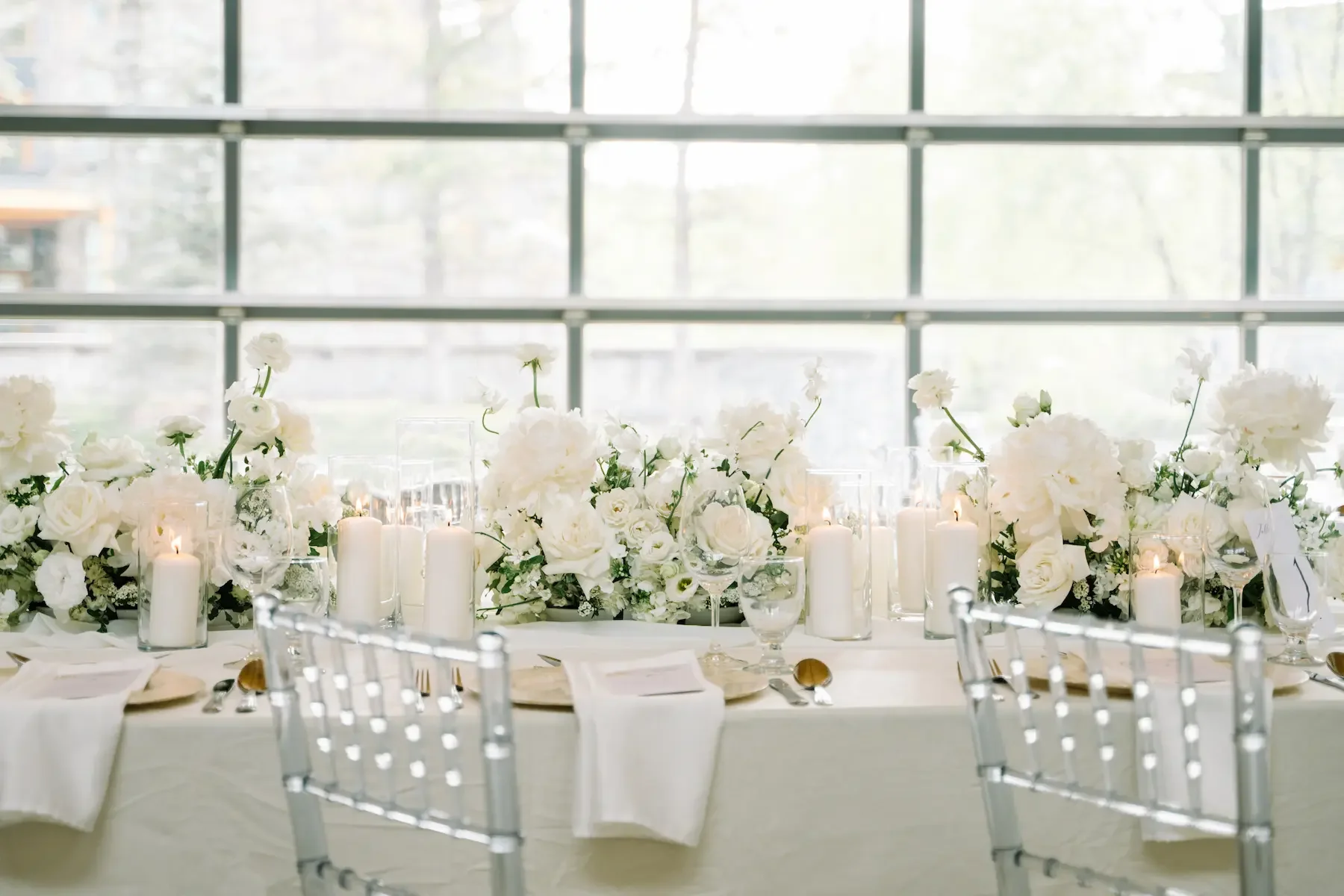 Elegant wedding table setting with white floral centerpiece and candles in glass holders, silver chairs, large windows in the background.