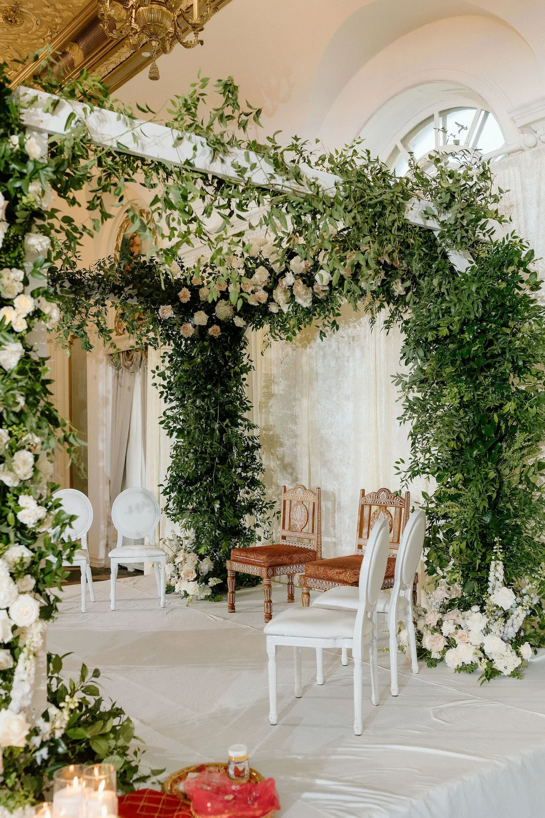 Elegant indoor wedding ceremony setup with floral arch, greenery, and chairs for the bride and groom.