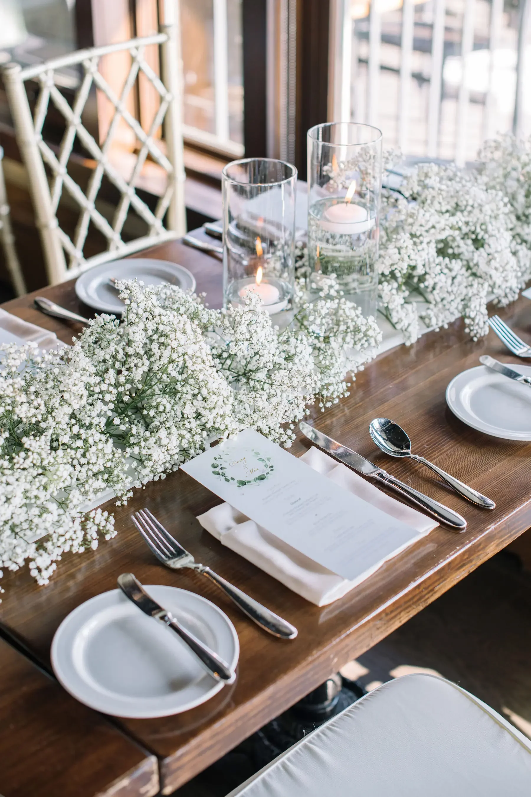 Elegant dining table set with white floral runner, candles in glass holders, white plates, silverware, and a printed menu, near a window with sunlight.
