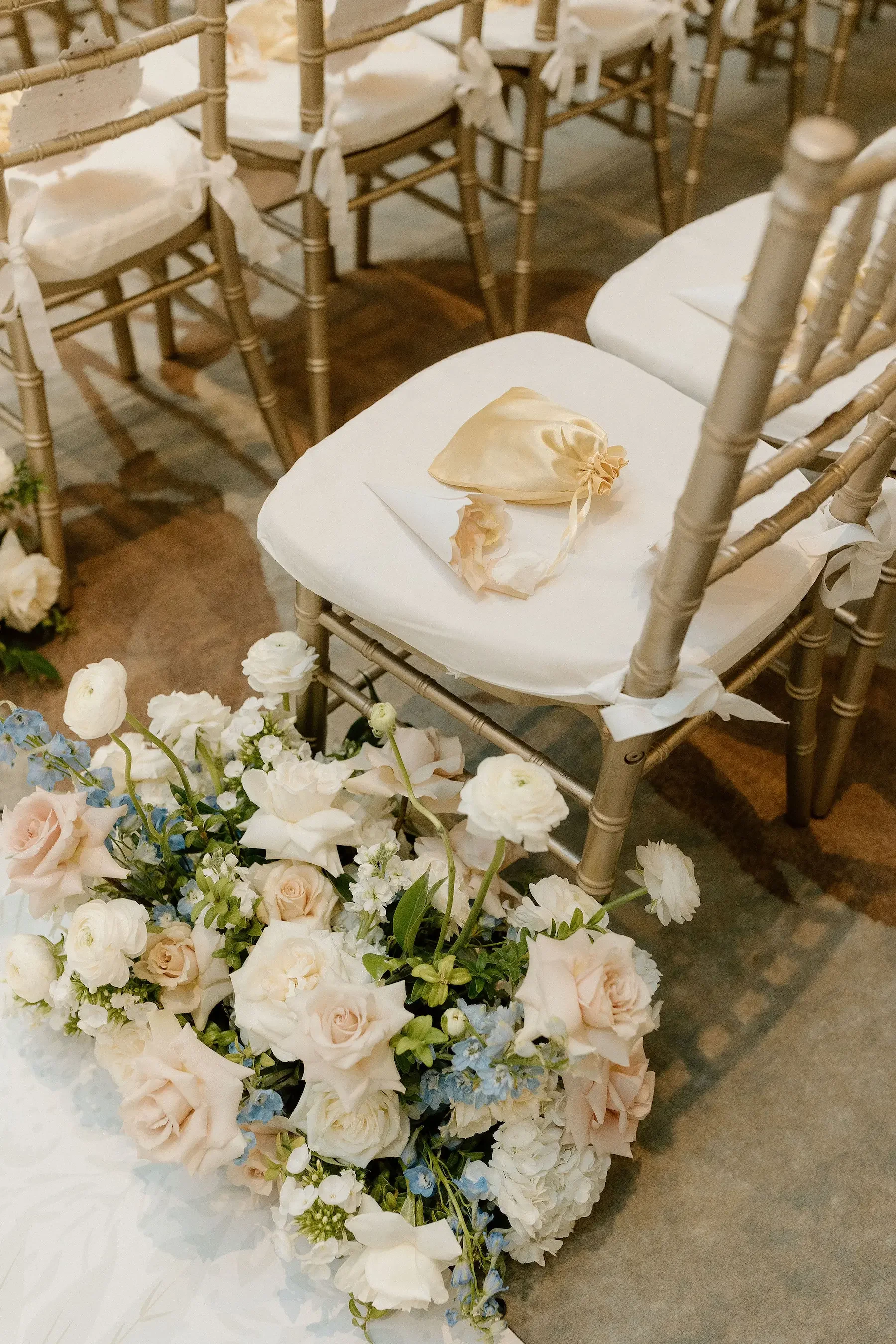 Empty white chairs with white cushions and bows, surrounded by a large floral arrangement of white, blush, and blue flowers for a wedding or special event.