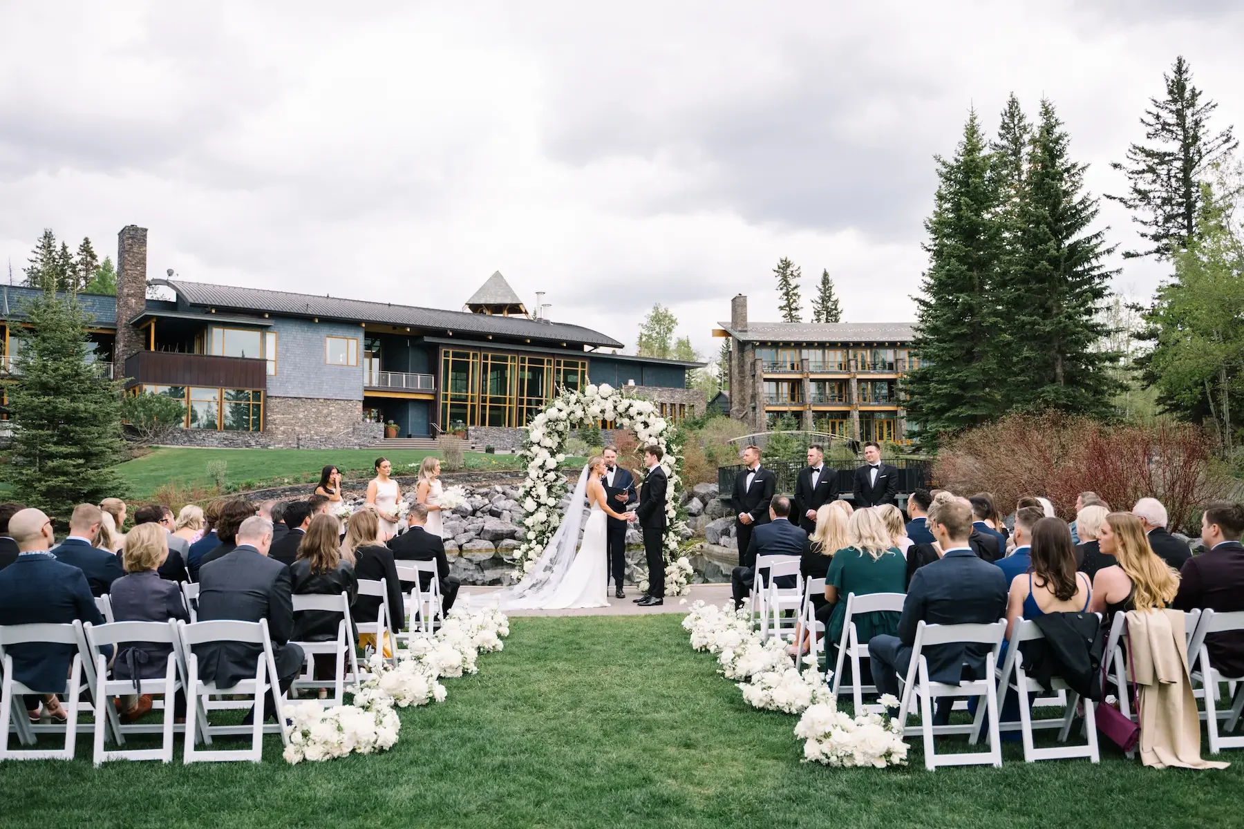 An outdoor wedding ceremony with a bride and groom standing under a floral arch, surrounded by seated guests on a grassy area, with a scenic backdrop of trees, rocks, and modern buildings.
